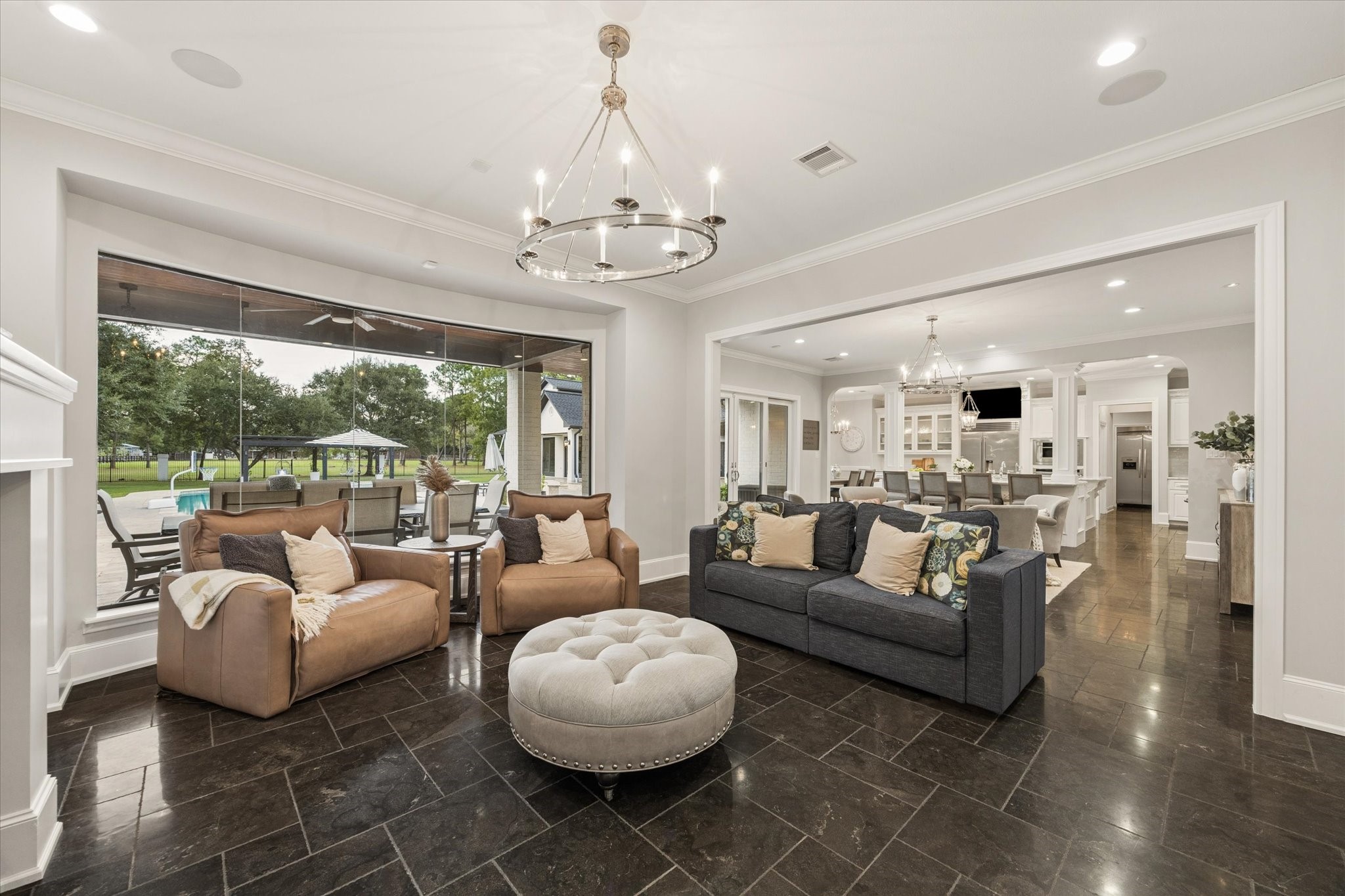 Spacious living area with elegant lighting, featuring large windows offering views of the lush backyard and pool. The open-concept design flows seamlessly into a modern kitchen, perfect for entertaining.