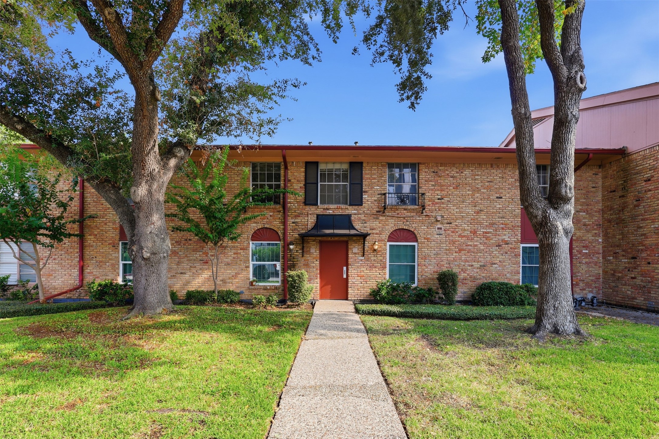 Charming brick facade welcomes you to this inviting entrance, framed by mature trees and lush greenery, offering a perfect blend of elegance and warmth.
