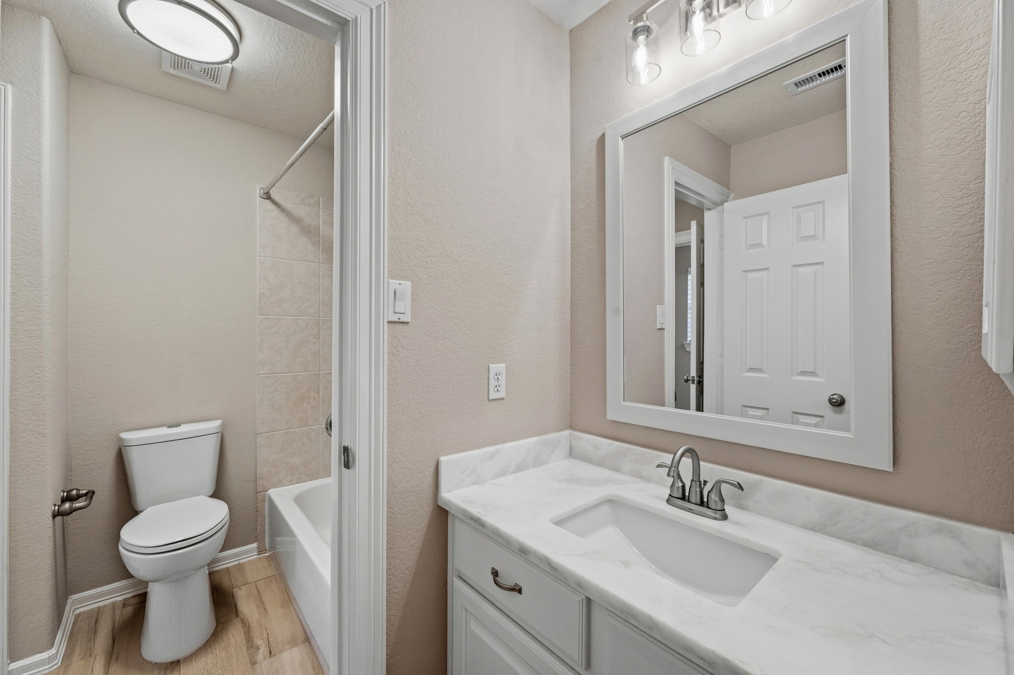 Charming bathroom featuring a sleek vanity, modern lighting, and a classic tub-shower combination, all complemented by warm wood-look flooring.
