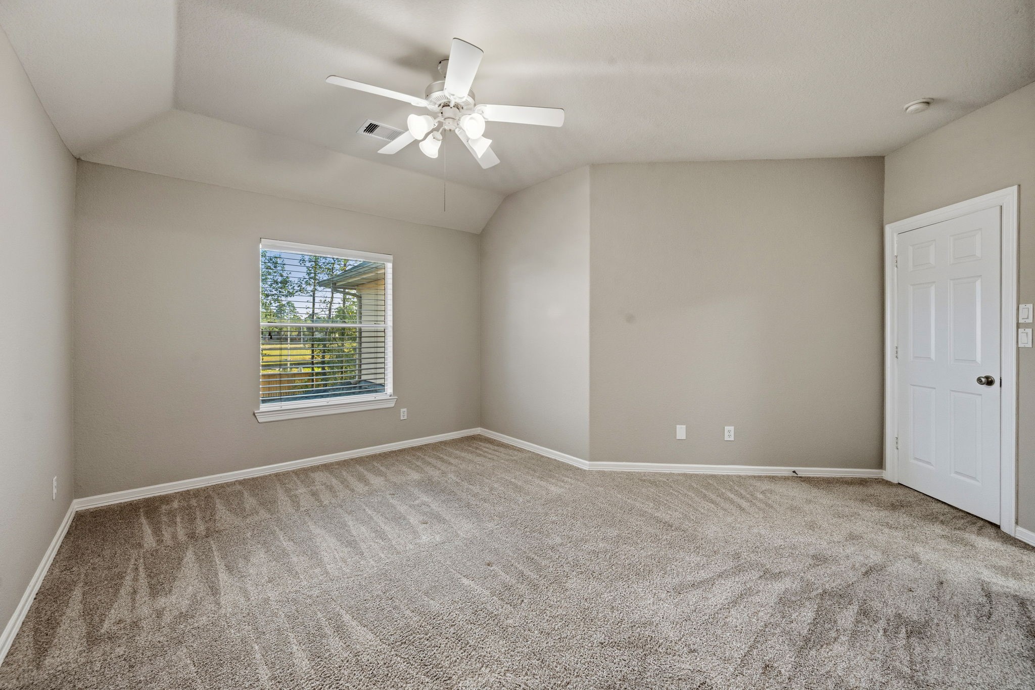 Spacious bedroom with plush carpeting, neutral walls, and ample natural light from a large window, complemented by a ceiling fan for comfort.