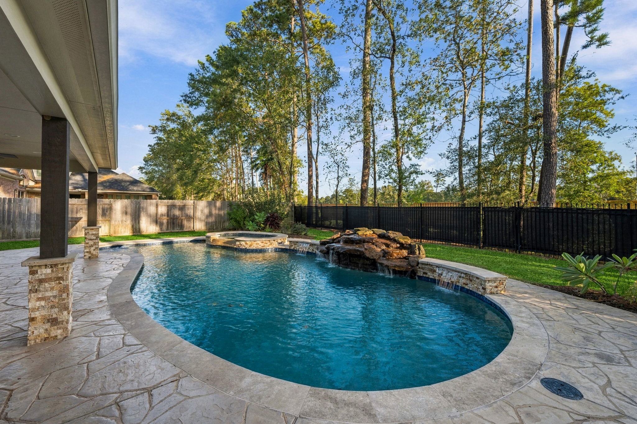 Stunning backyard oasis featuring a luxurious pool with a waterfall feature, framed by lush greenery and a spacious stone patio perfect for relaxation and entertaining.