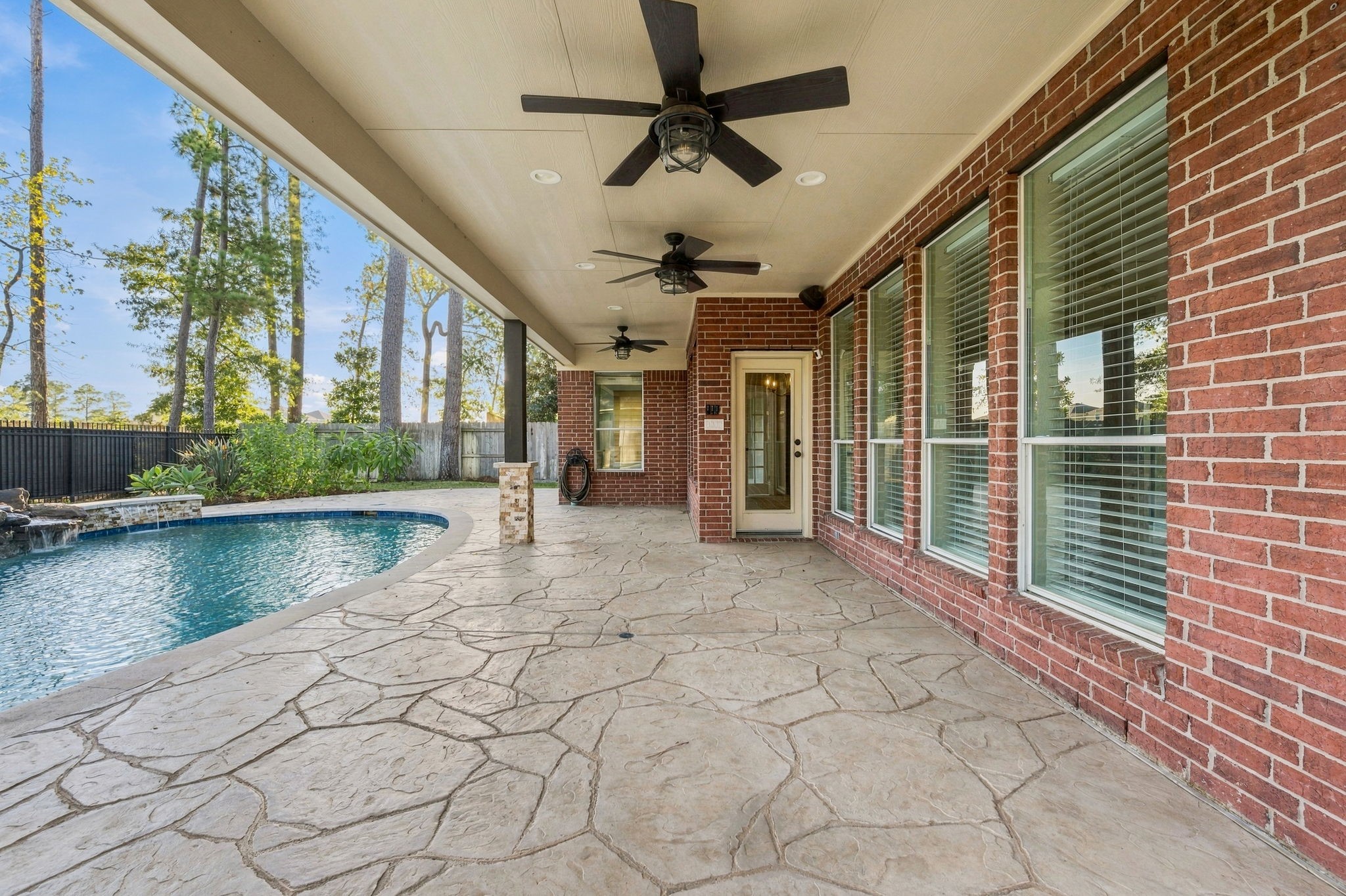 Spacious covered patio with elegant stone flooring and ceiling fans, overlooking a sparkling pool set amid a serene backdrop.