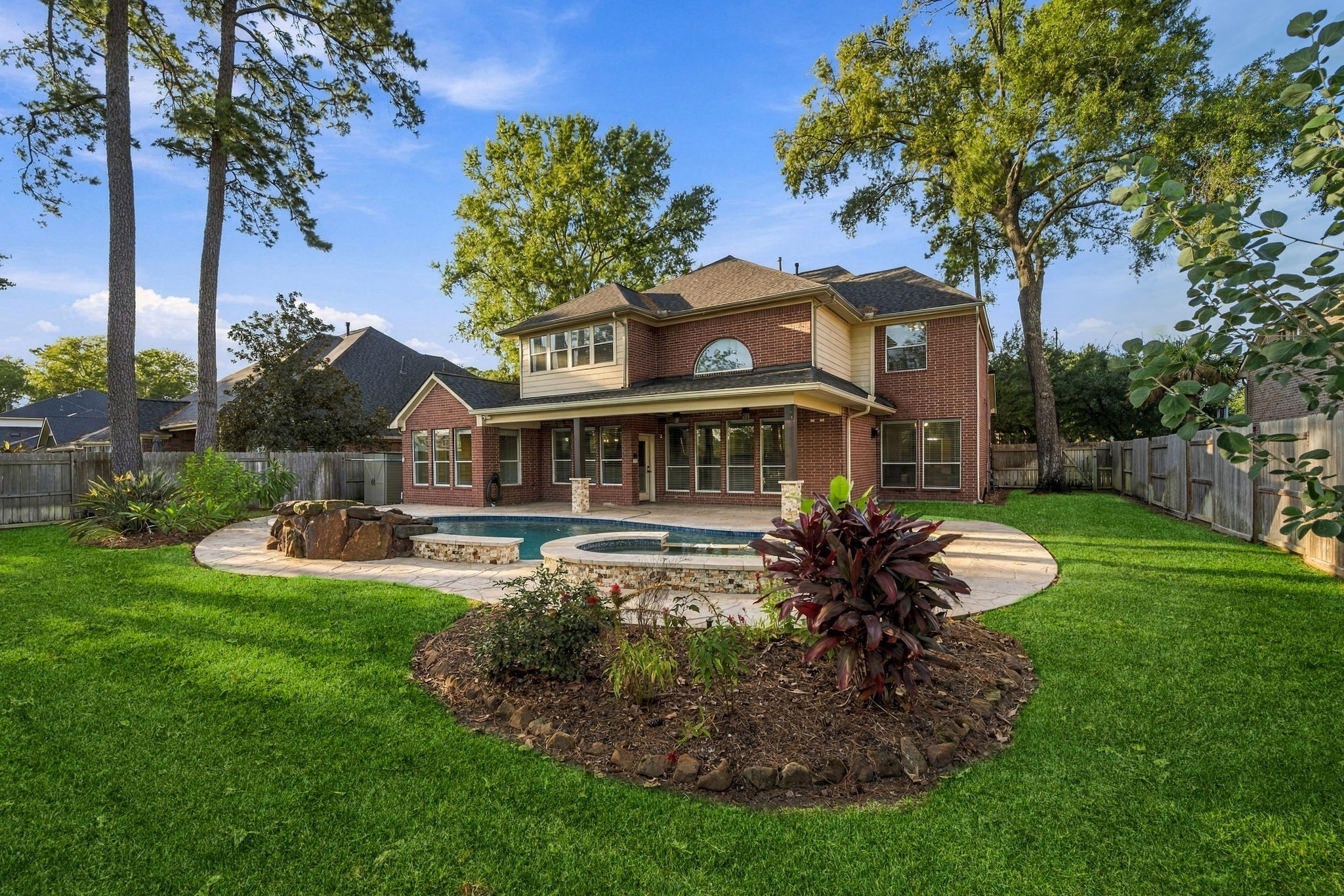 Spacious backyard featuring a sparkling pool, lush landscaping, and a charming brick home with large windows and a covered patio, perfect for outdoor entertaining.