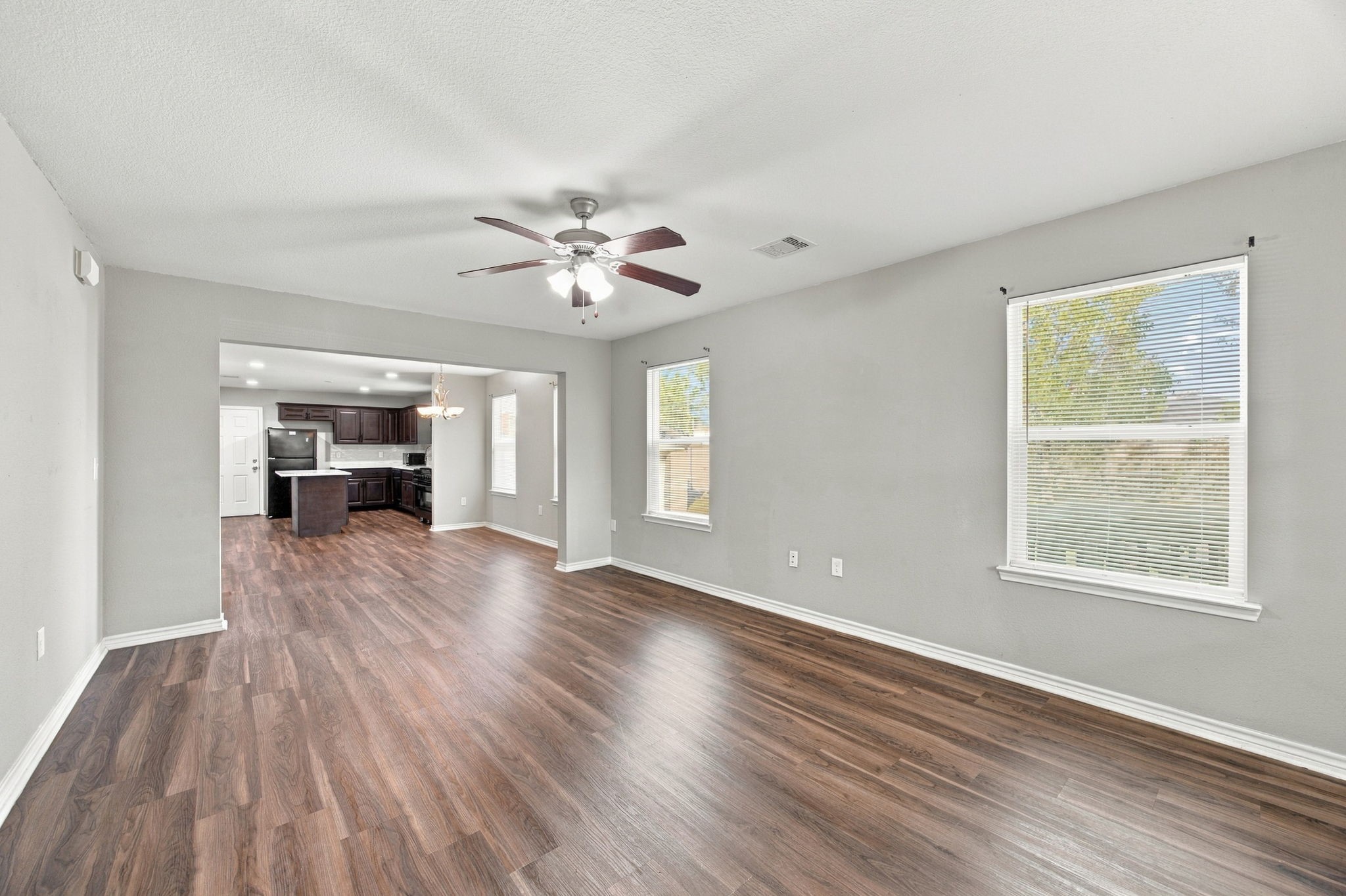 Spacious living area featuring wood-look flooring, neutral walls, a ceiling fan, and an open connection to the kitchen.