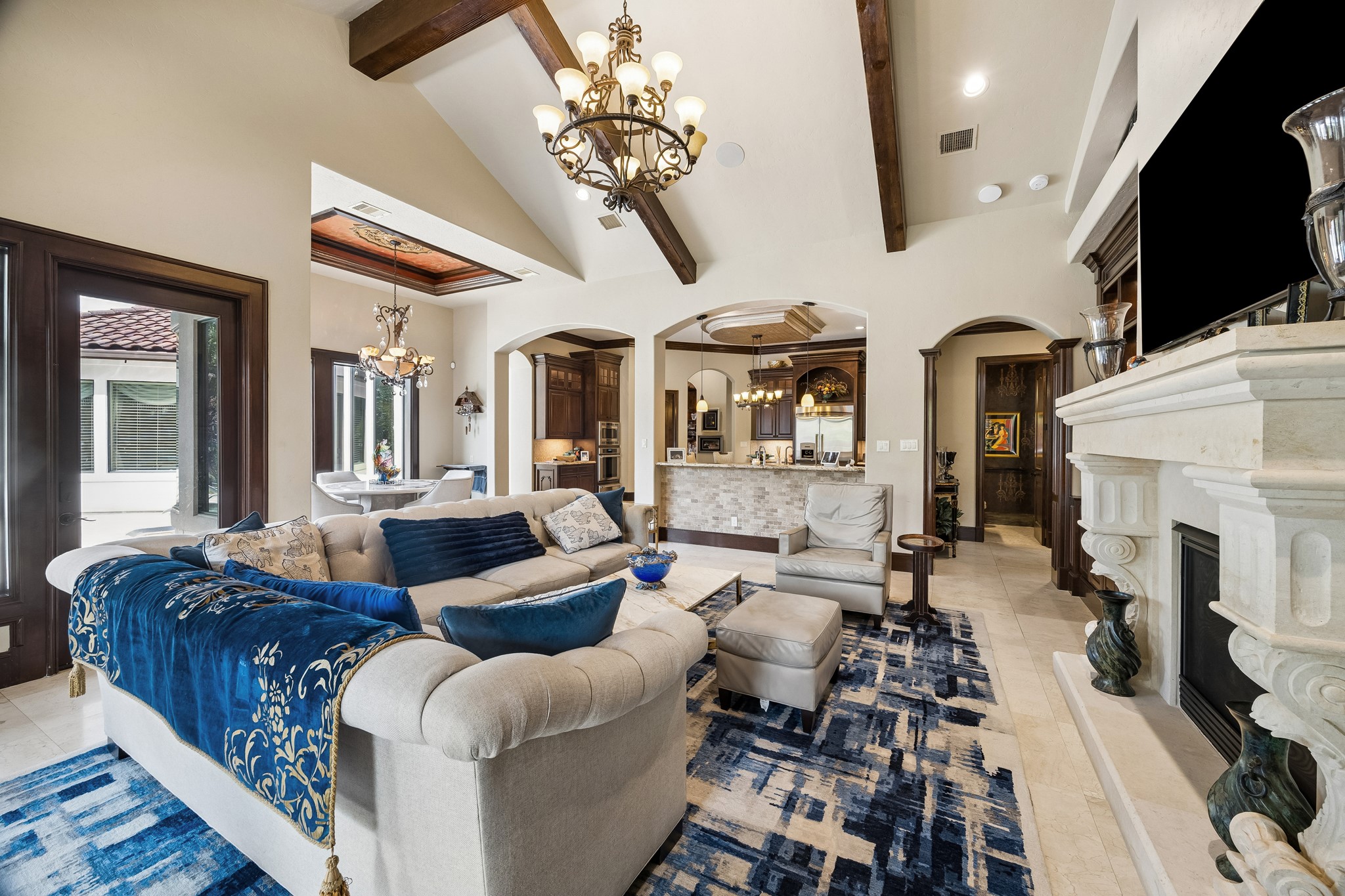 Elegant living space featuring high ceilings with exposed beams, a grand stone fireplace, and luxurious chandeliers. The open layout connects seamlessly to a sophisticated kitchen, perfect for entertaining.