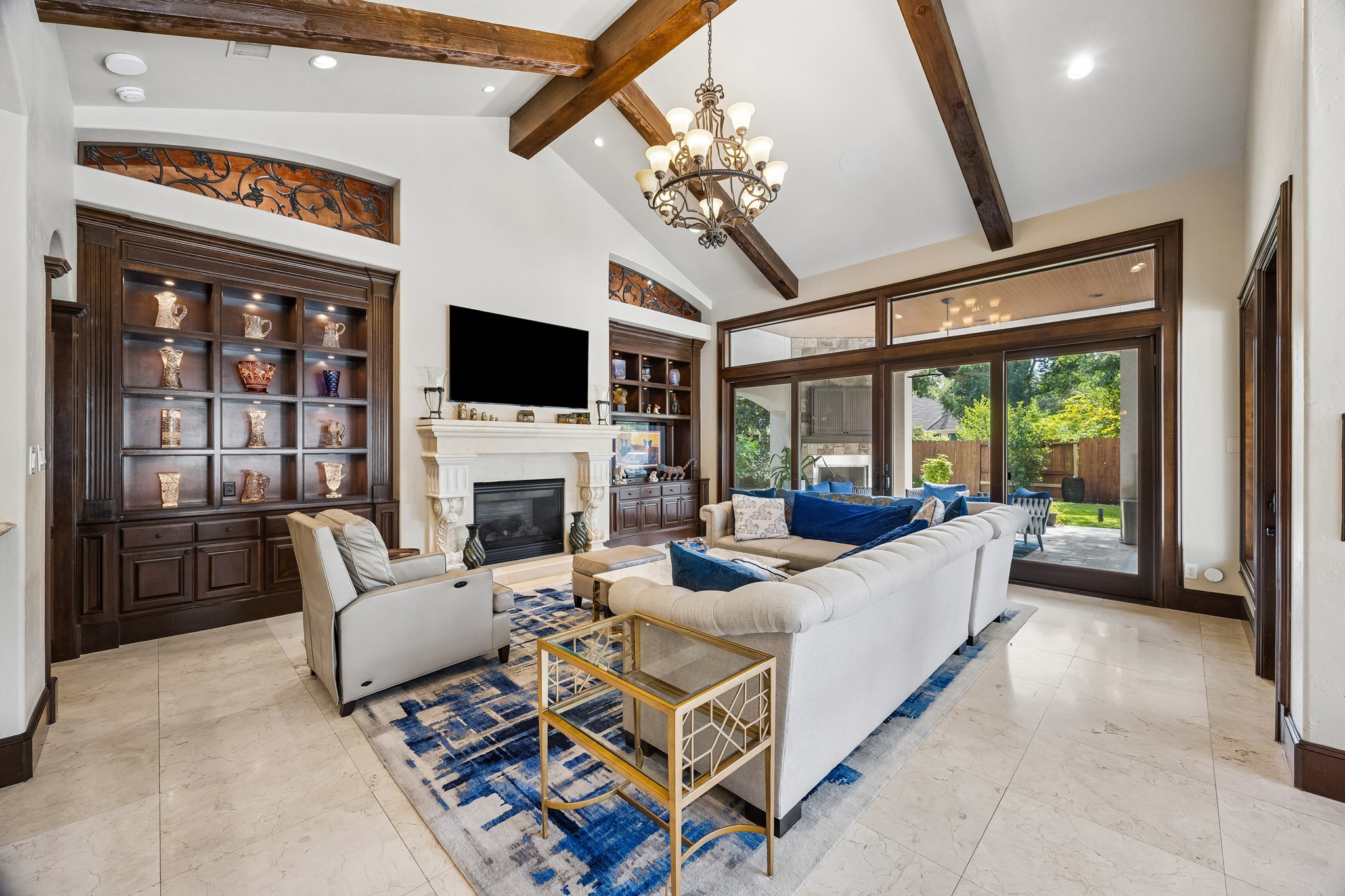 Spacious family room with elegant wooden beams, a grand fireplace, and large windows offering a view of the lush backyard. There is a covered patio with a fireplace and summer kitchen just off the family room.