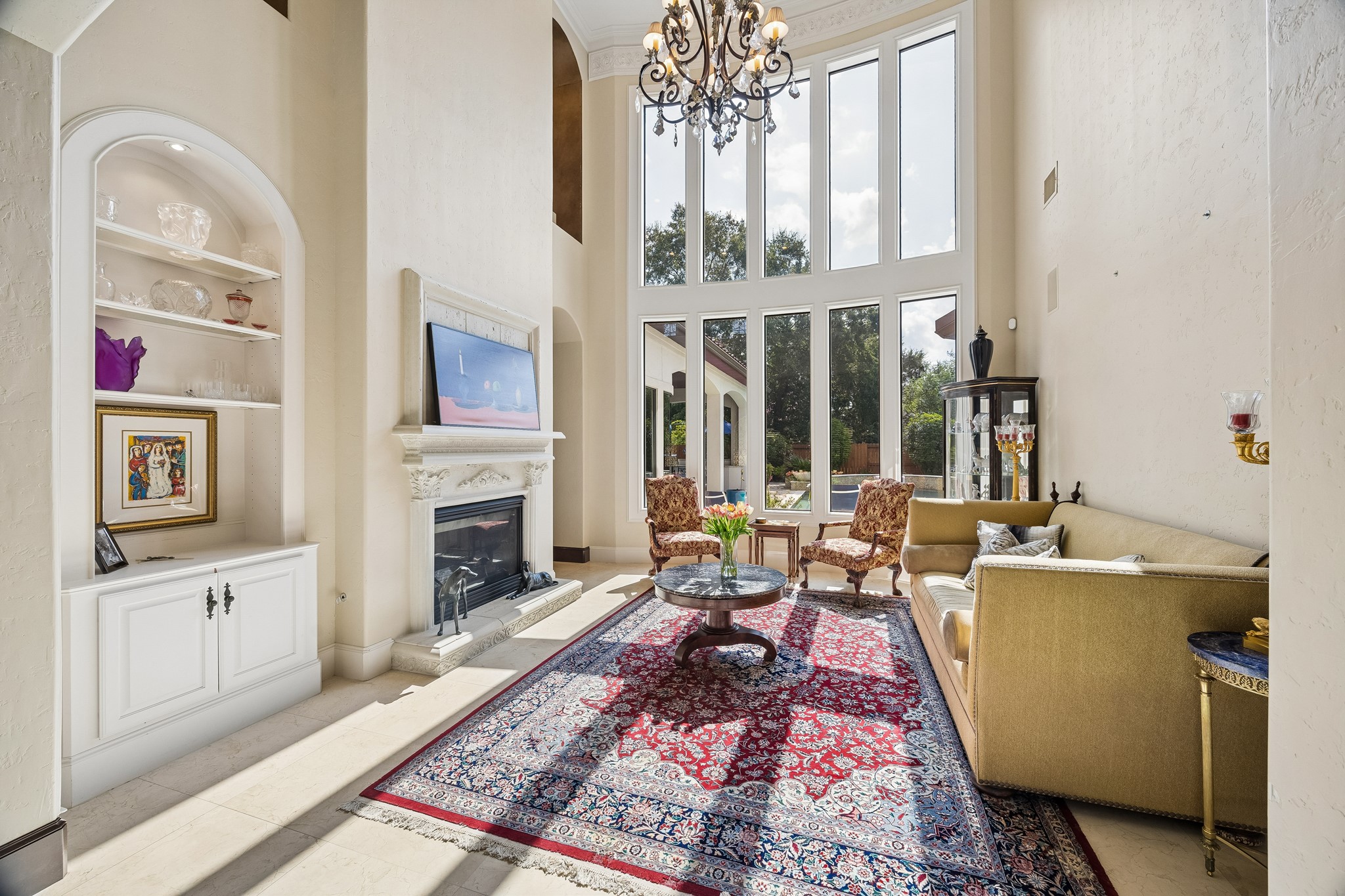 Grand living room with soaring ceilings, elegant chandelier, and a wall of windows that flood the space with natural light. Cozy fireplace and built-in shelving add charm, while the plush seating area offers a perfect spot for relaxation.