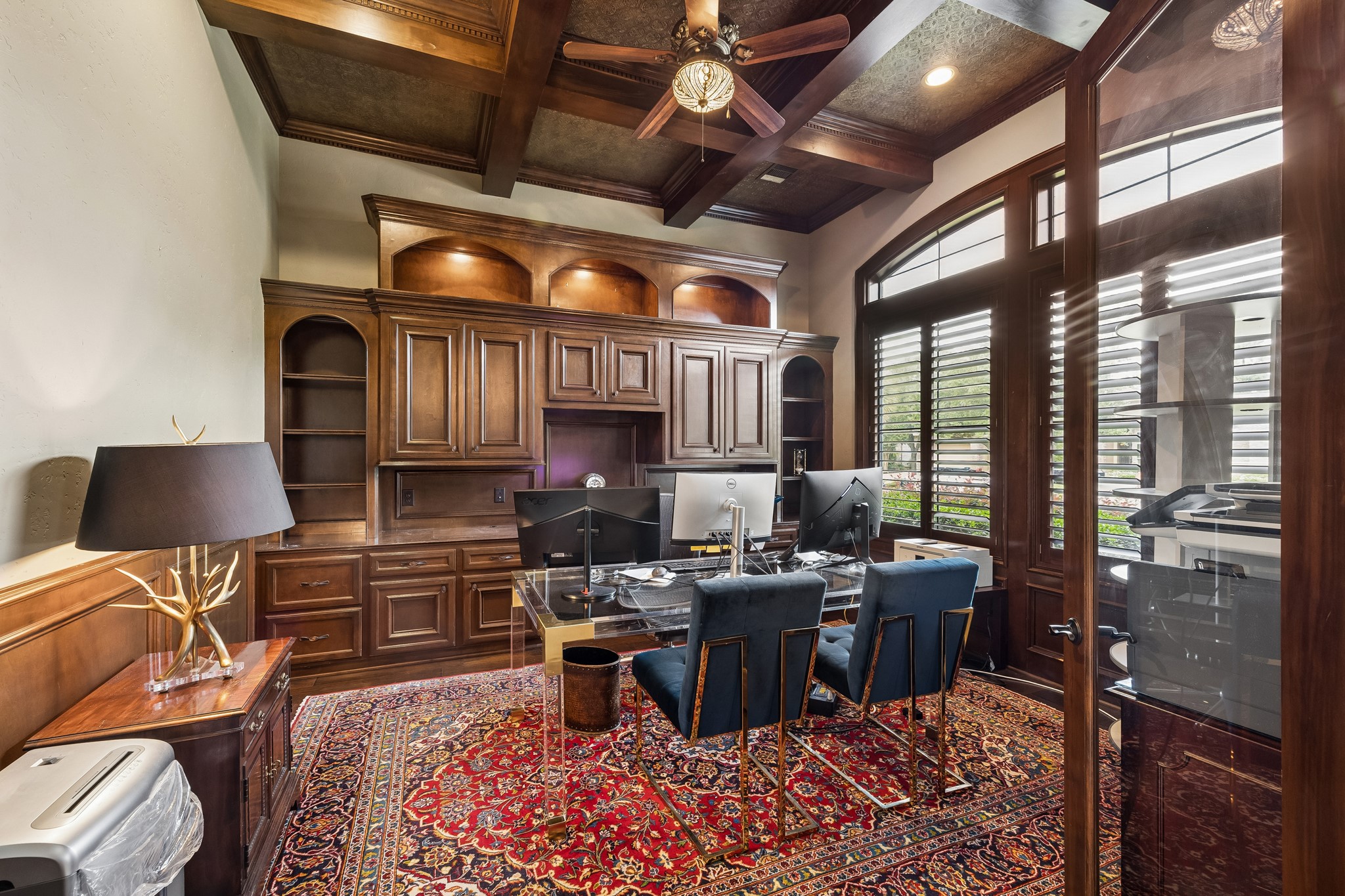 Elegant library featuring rich wood cabinetry, a coffered ceiling, and large windows with plantation shutters.
