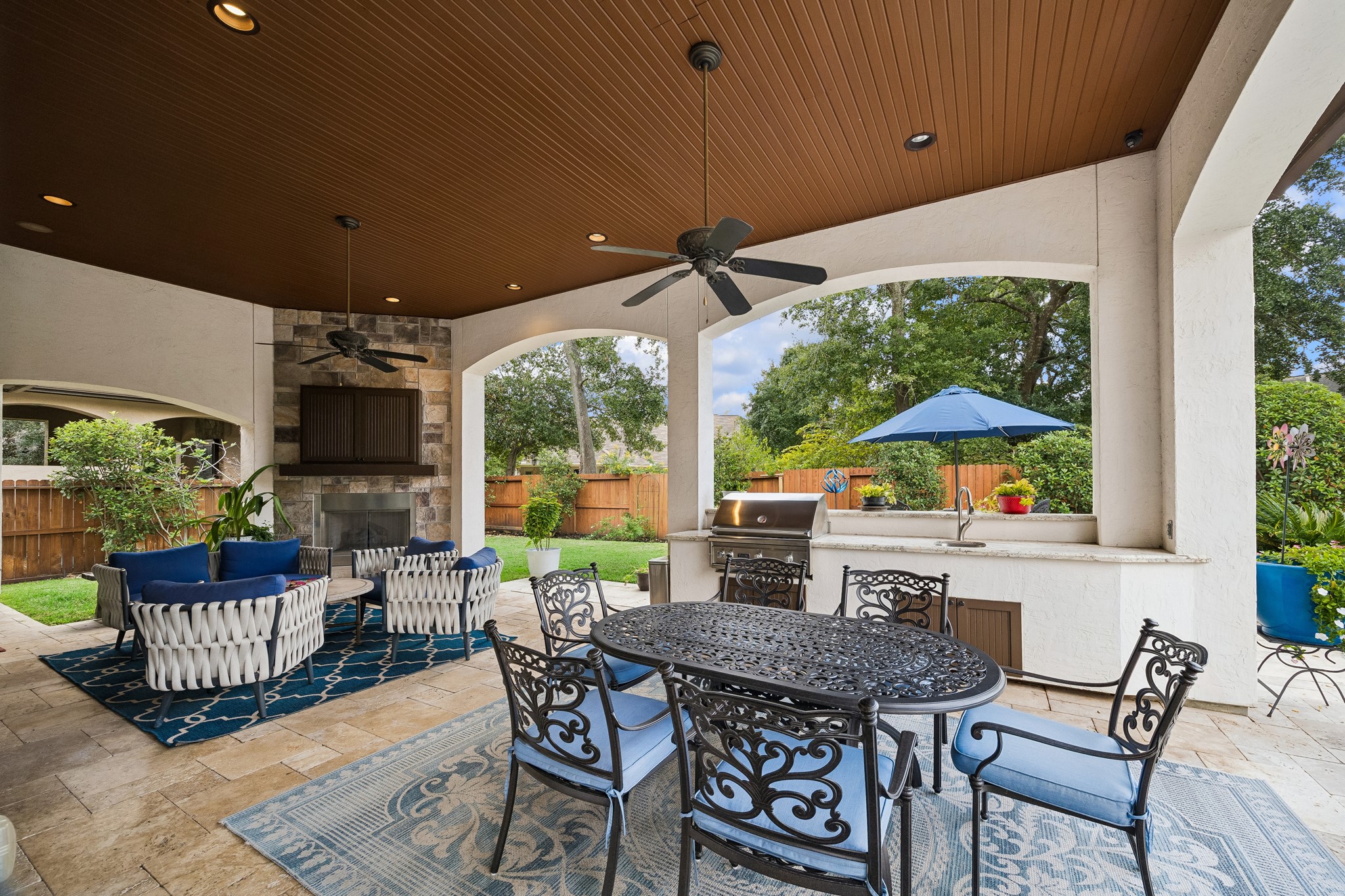 Luxurious outdoor living space featuring a covered patio with an outdoor TV, cozy seating around a fireplace, plus additional seating for outdoor dining, Viking built-in grill, and lush garden views.