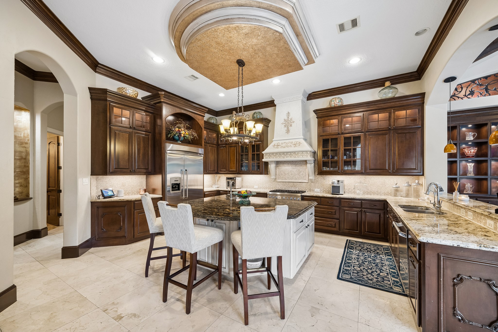 Elegant kitchen featuring rich wooden cabinetry, a large central island with seating, and high-end stainless steel appliances including a built-in fridge/freezer, 6 burner gas range with professional vent hood, microwave, dishwasher and warming drawer all under a stunning coffered ceiling.