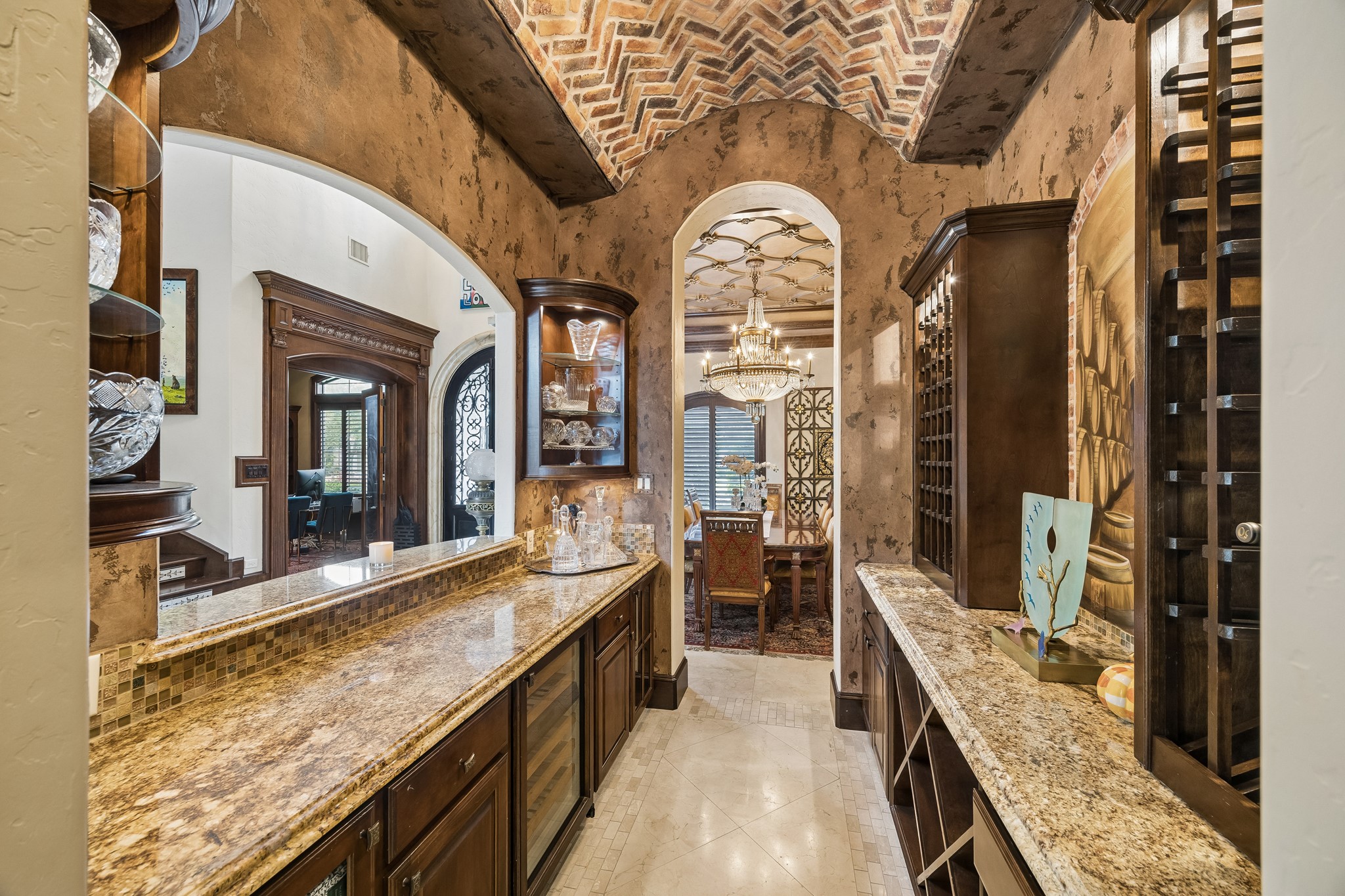 Step into the luxurious bar featuring granite counters, rich wood cabinetry, and a stunning brick barrel vaulted ceiling, perfectly situated off the grand entrance and dining area adorned with a sparkling chandelier.