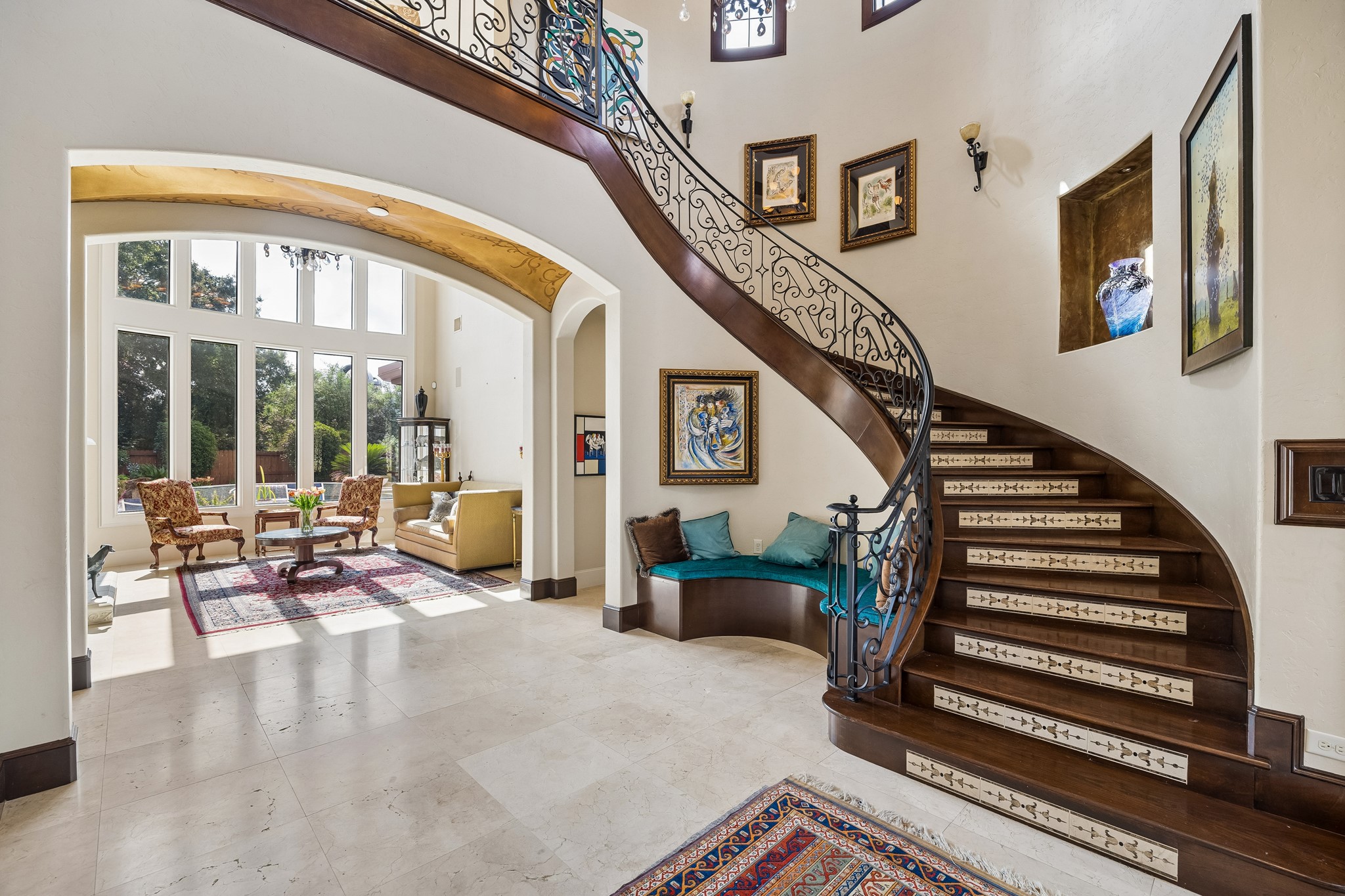 Grand foyer with a stunning curved staircase, ornate iron railing, and a cozy seating nook. Abundant natural light floods the adjacent living room, creating a welcoming atmosphere.