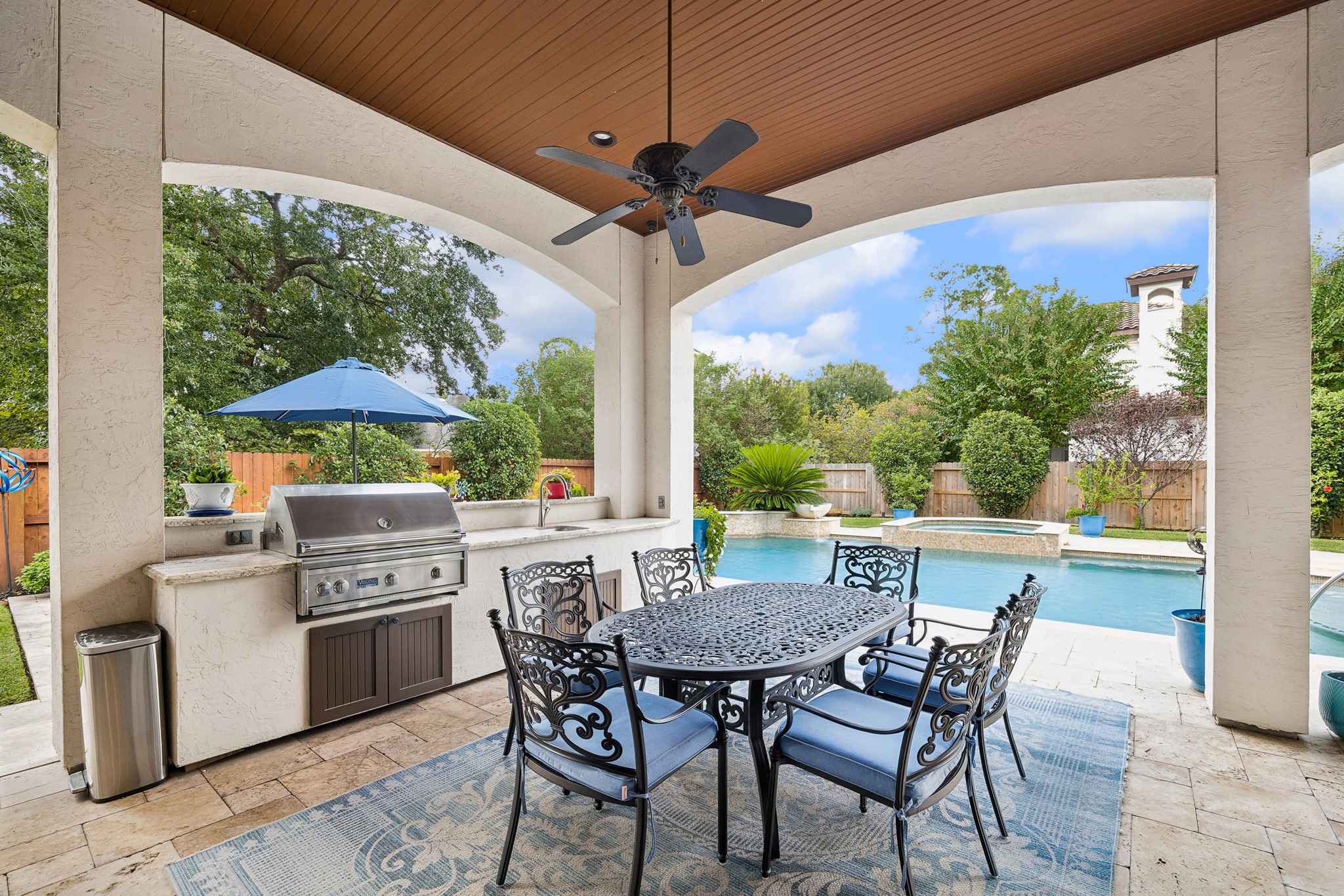 Experience luxurious outdoor living with a covered patio featuring an elegant dining area, built-in Viking grill, and views of a sparkling pool surrounded by lush landscaping.