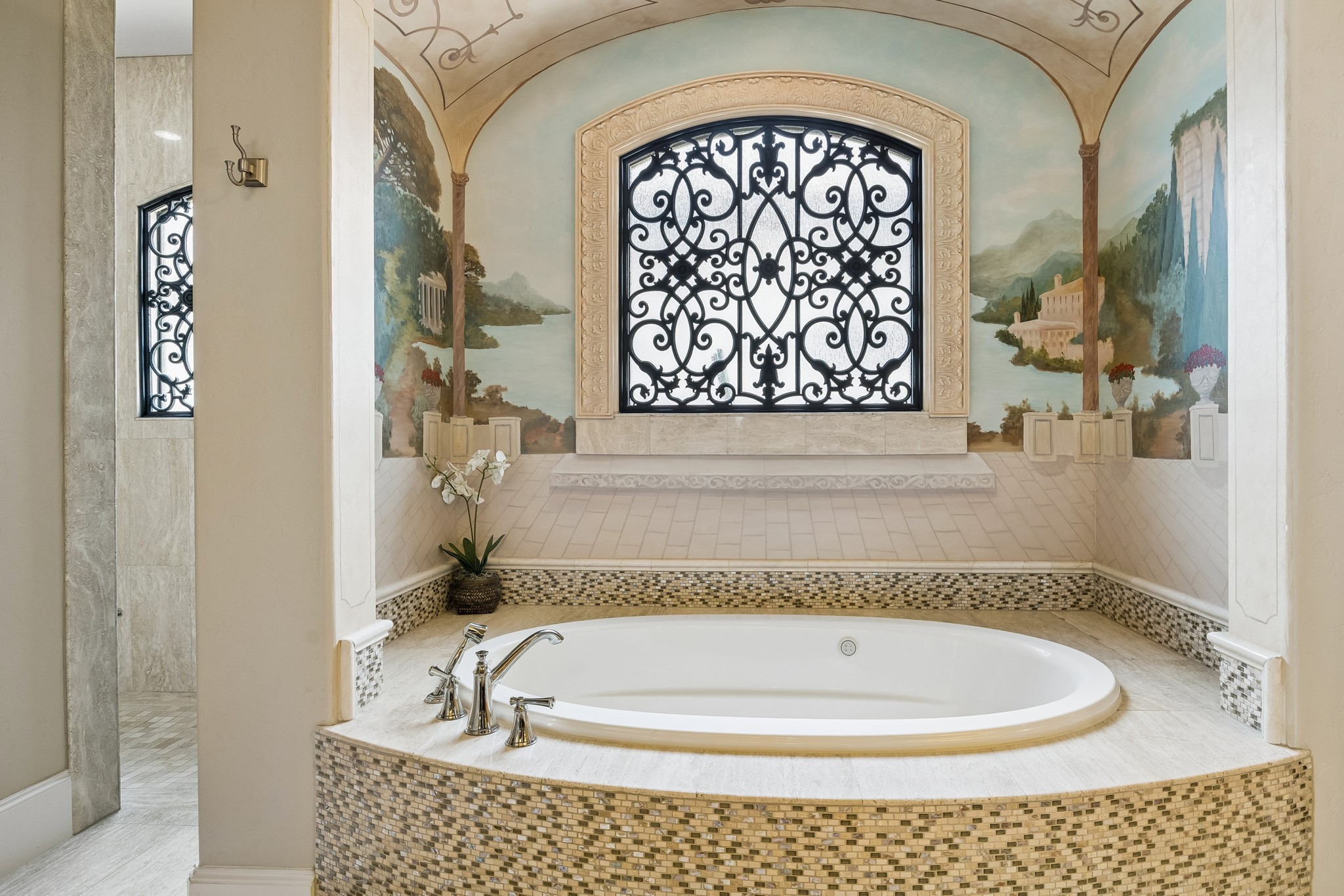 Luxurious bathroom featuring a stunning soaking tub with elegant mosaic tiling, set beneath a beautifully detailed mural and decorative arched window, creating a serene retreat.