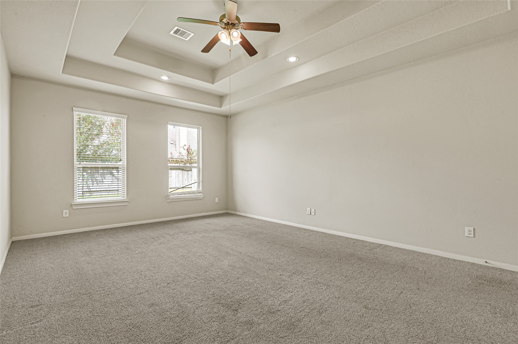 Large bedroom with ceiling fan, tray ceiling, multiple windows, and neutral carpet.