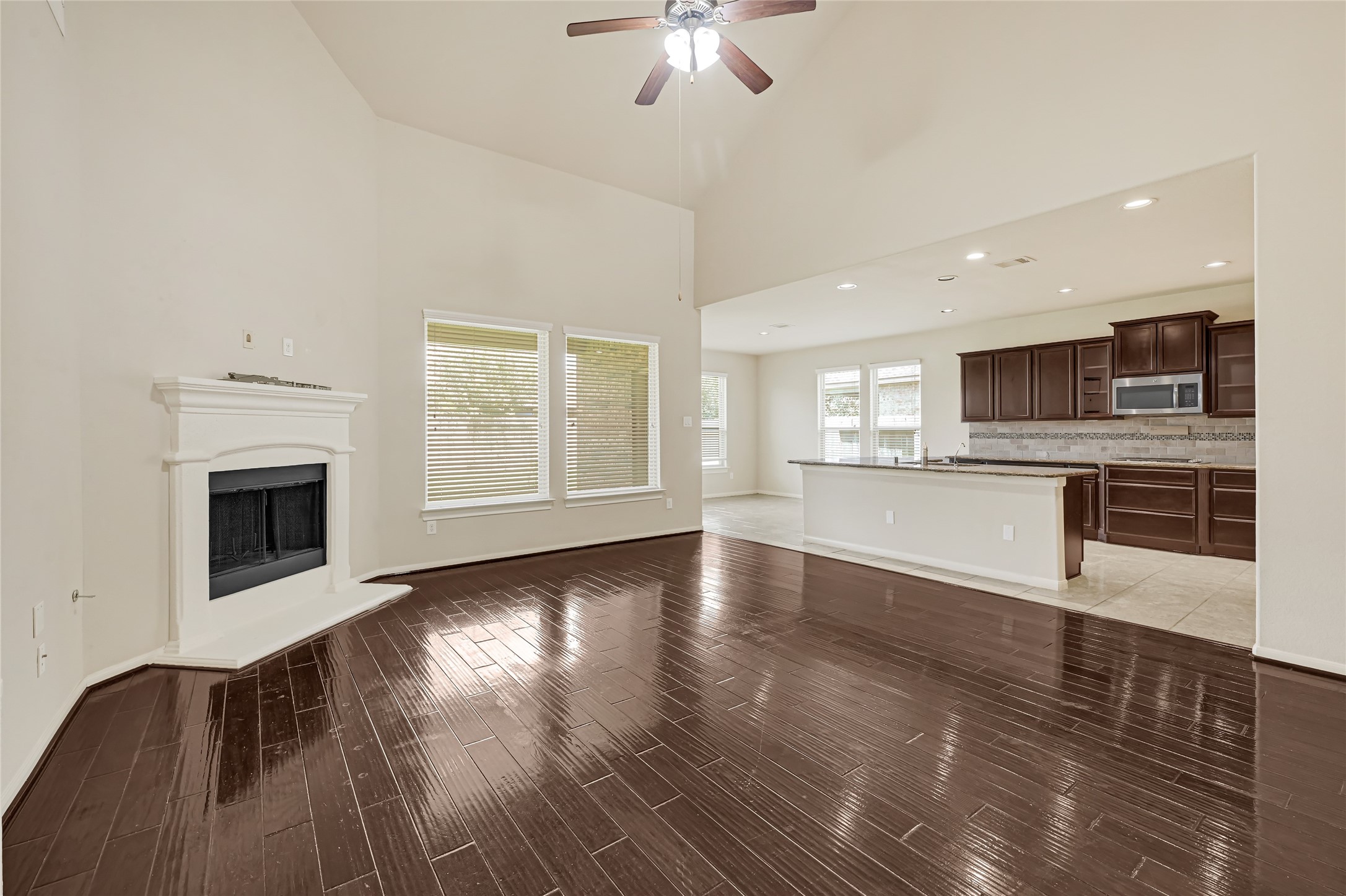 Spacious living room with hardwood flooring, a fireplace, ceiling fan, and views into the kitchen.