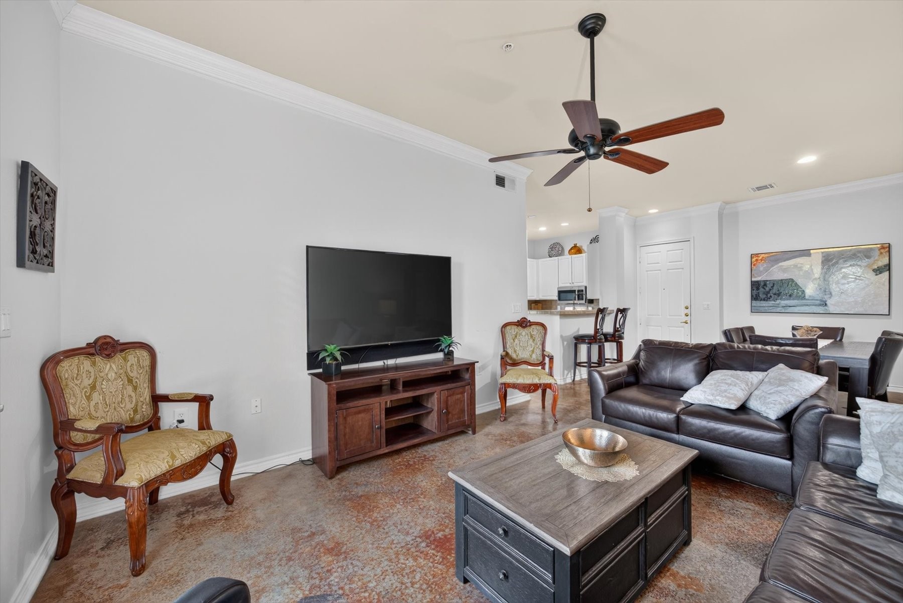 Spacious living area with elegant crown molding, polished flooring, and a large ceiling fan. The open layout features a comfortable seating arrangement, a wall-mounted TV, and a seamless flow into the dining and kitchen areas, perfect for entertaining.