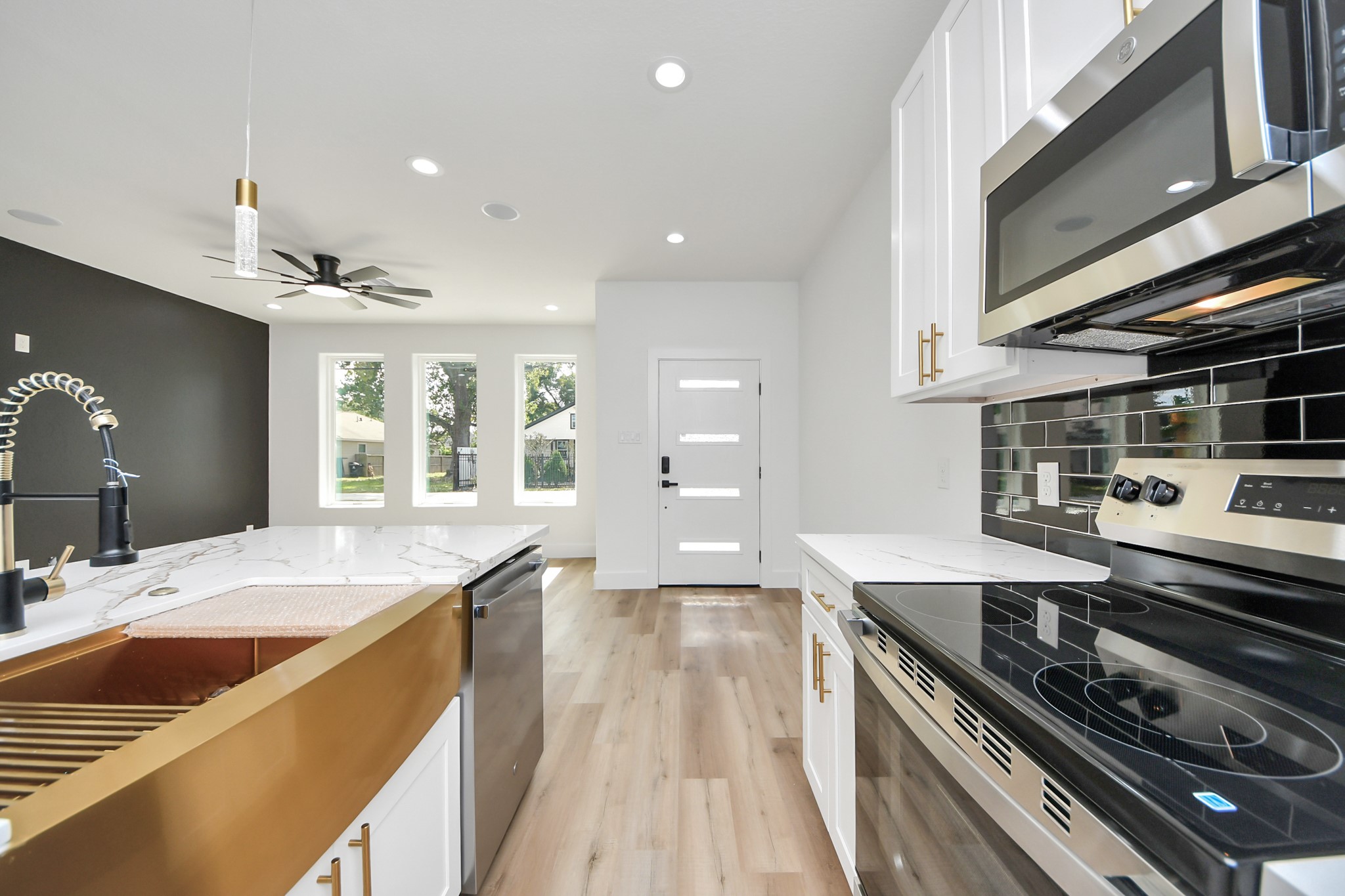Modern kitchen with sleek stainless steel appliances, elegant black backsplash, and a stylish gold-accented sink, seamlessly flowing into a bright living area with large windows and contemporary ceiling fan.