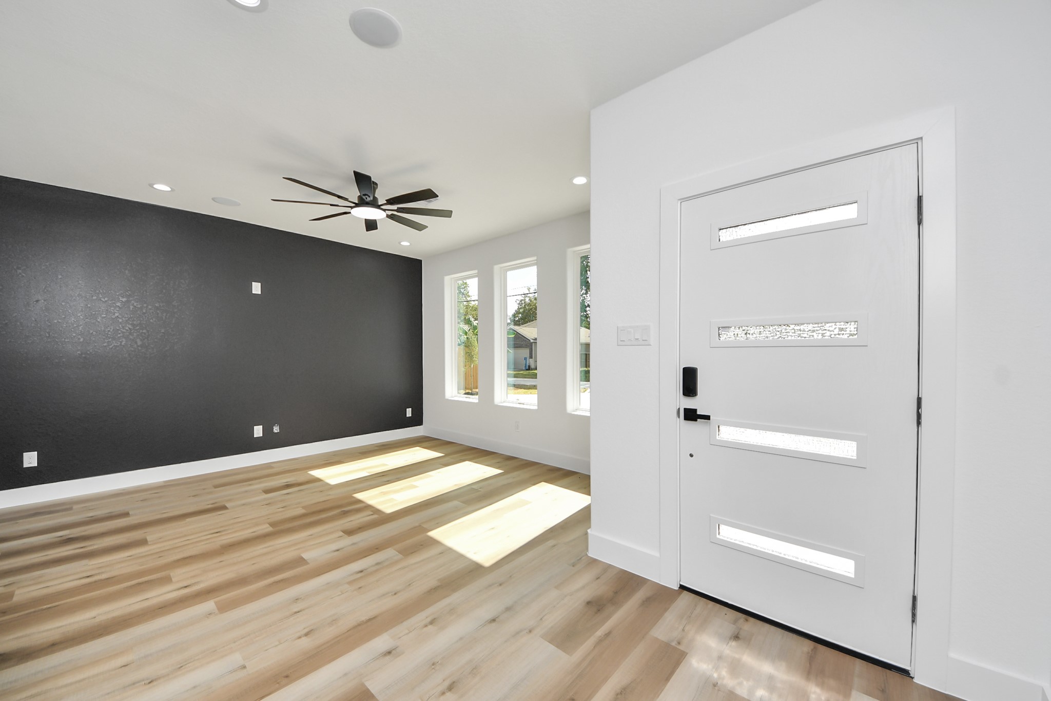 Bright and modern entryway featuring a sleek front door, large windows, wood flooring, and a bold accent wall with a contemporary ceiling fan.