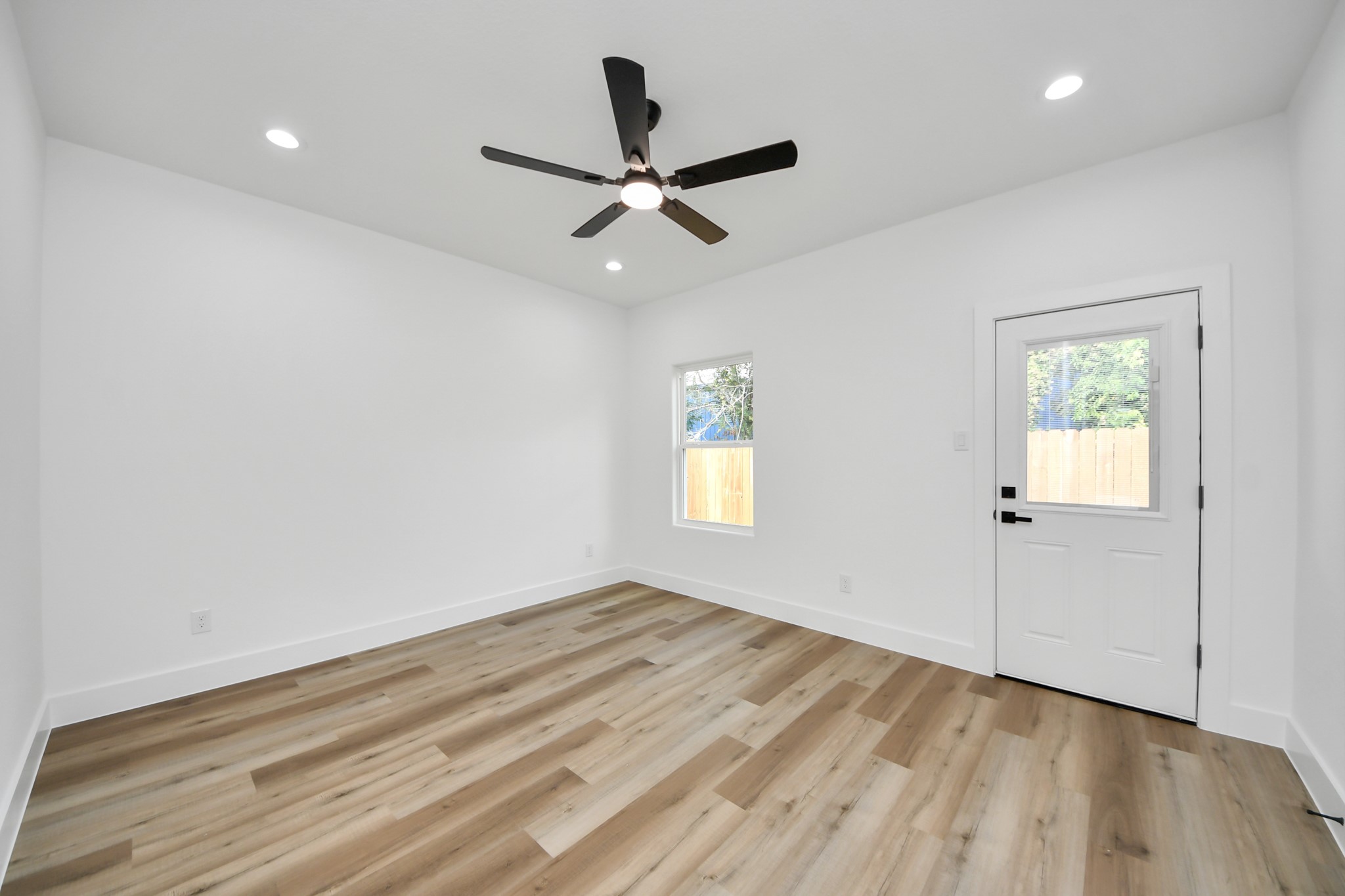 Bright and inviting room featuring sleek hardwood floors, recessed lighting, and a modern ceiling fan, complemented by a window and door for ample natural light.