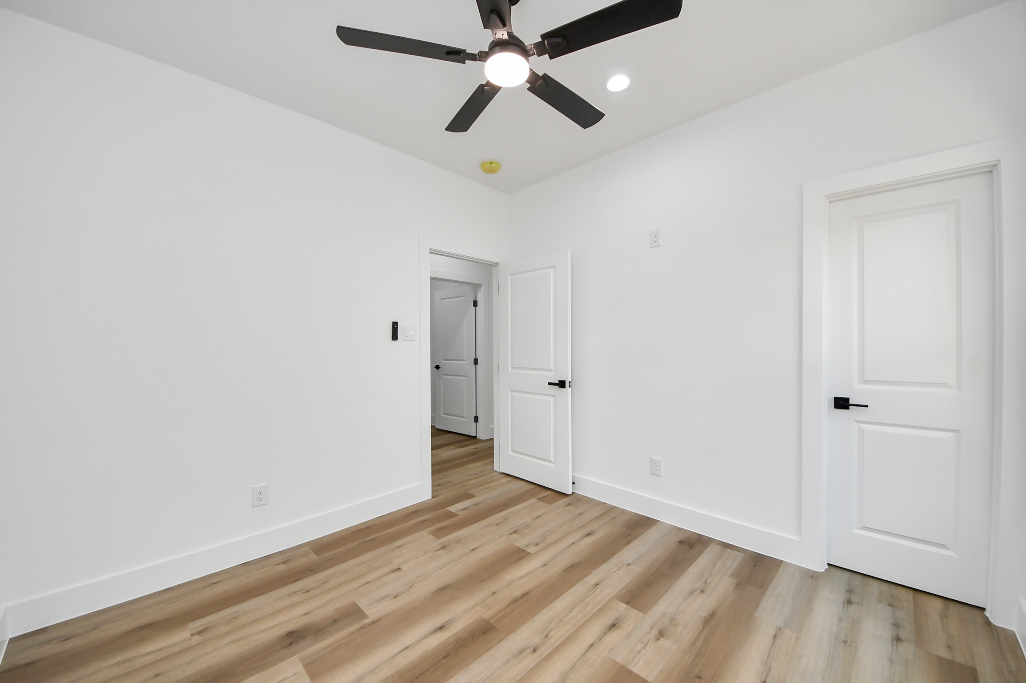 Bright and airy bedroom featuring sleek wood flooring, modern ceiling fan, and crisp white walls for a fresh, inviting atmosphere.