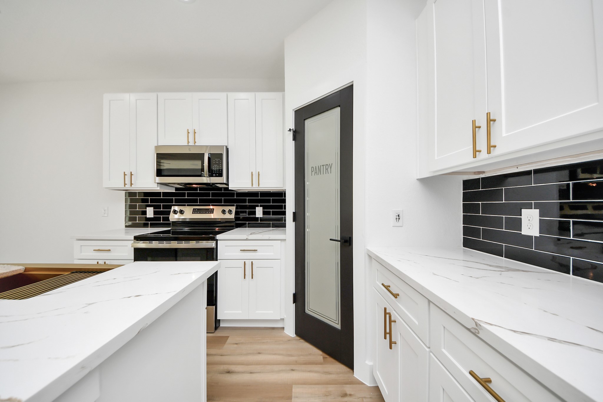 Modern kitchen featuring sleek white cabinetry, elegant marble-style countertops, and a striking black tile backsplash. Equipped with stainless steel appliances and a convenient pantry with a frosted glass door.