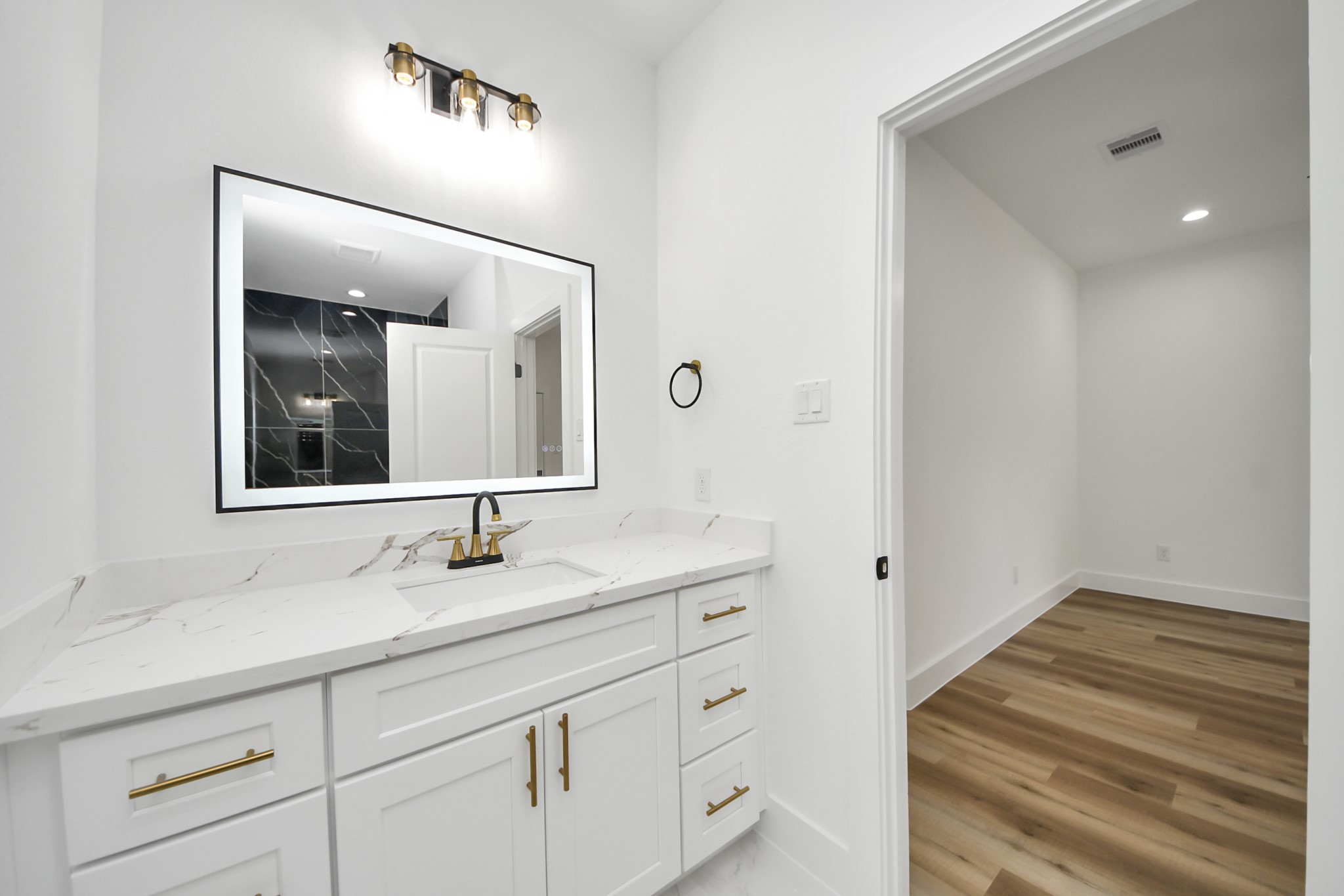 Elegant bathroom featuring a sleek vanity with a marble countertop, modern fixtures, and a large illuminated mirror, leading to a room with stylish wood flooring.