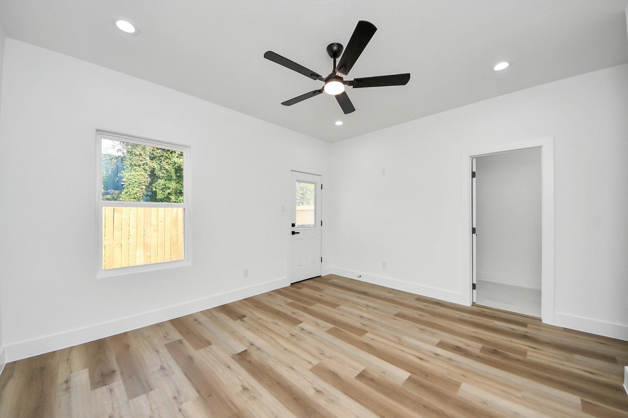 Bright and airy room featuring sleek hardwood floors, recessed lighting, and a modern ceiling fan, with a window and door leading outside for easy access.