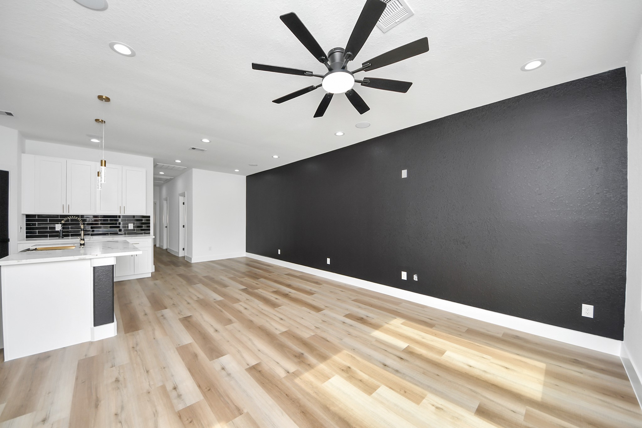 Spacious open-concept living area features sleek wood flooring and a striking black accent wall, seamlessly flowing into a modern kitchen with chic black-and-white cabinetry and stylish backsplash.
