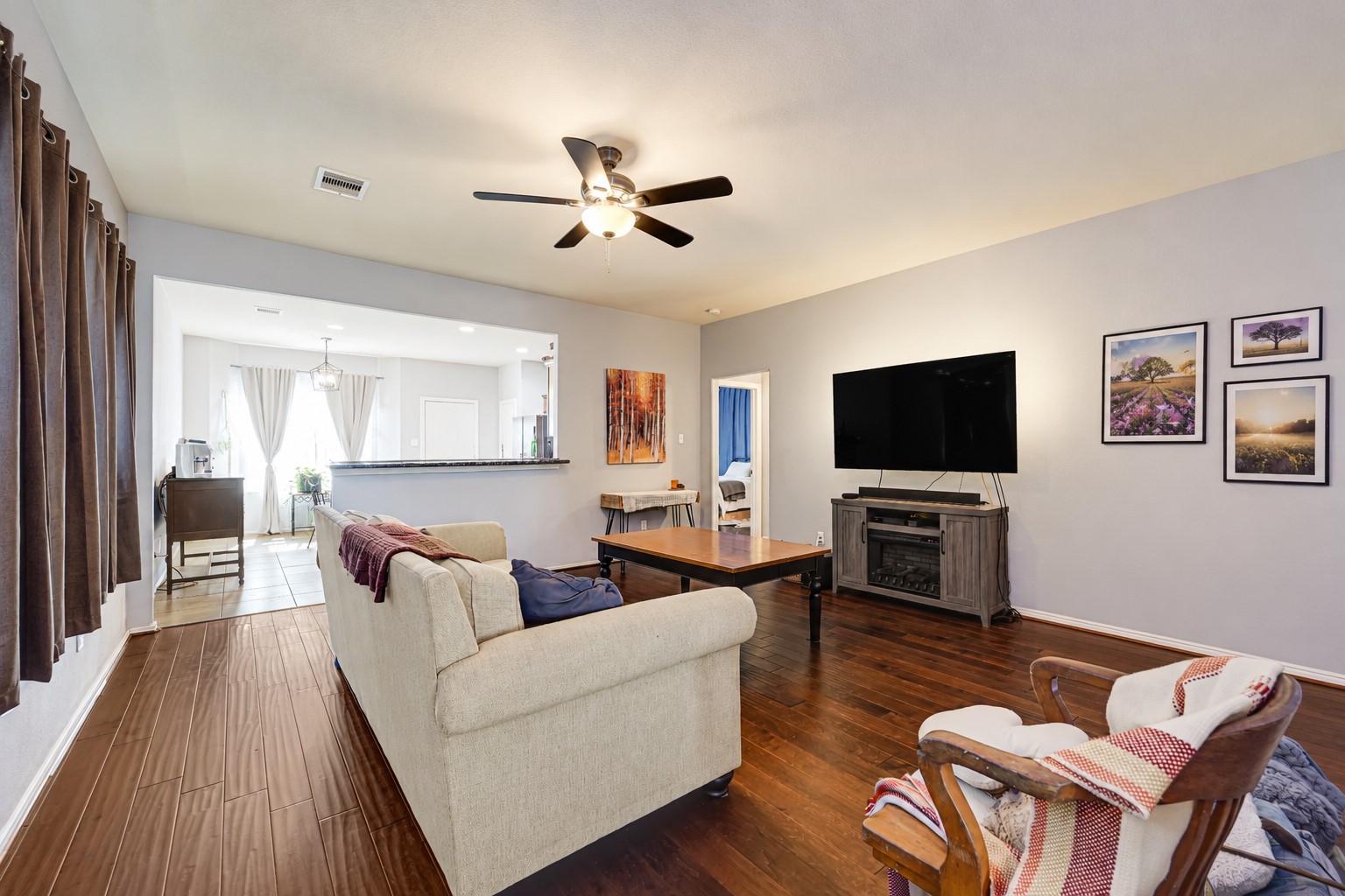Spacious living room featuring rich hardwood floors, a cozy fireplace, and a large wall-mounted TV, seamlessly flowing into a bright dining area with elegant drapes. Spacious living room featuring rich hardwood floors, a cozy fireplace, and a large wall-mounted TV, seamlessly flowing into a bright dining area with elegant drapes.