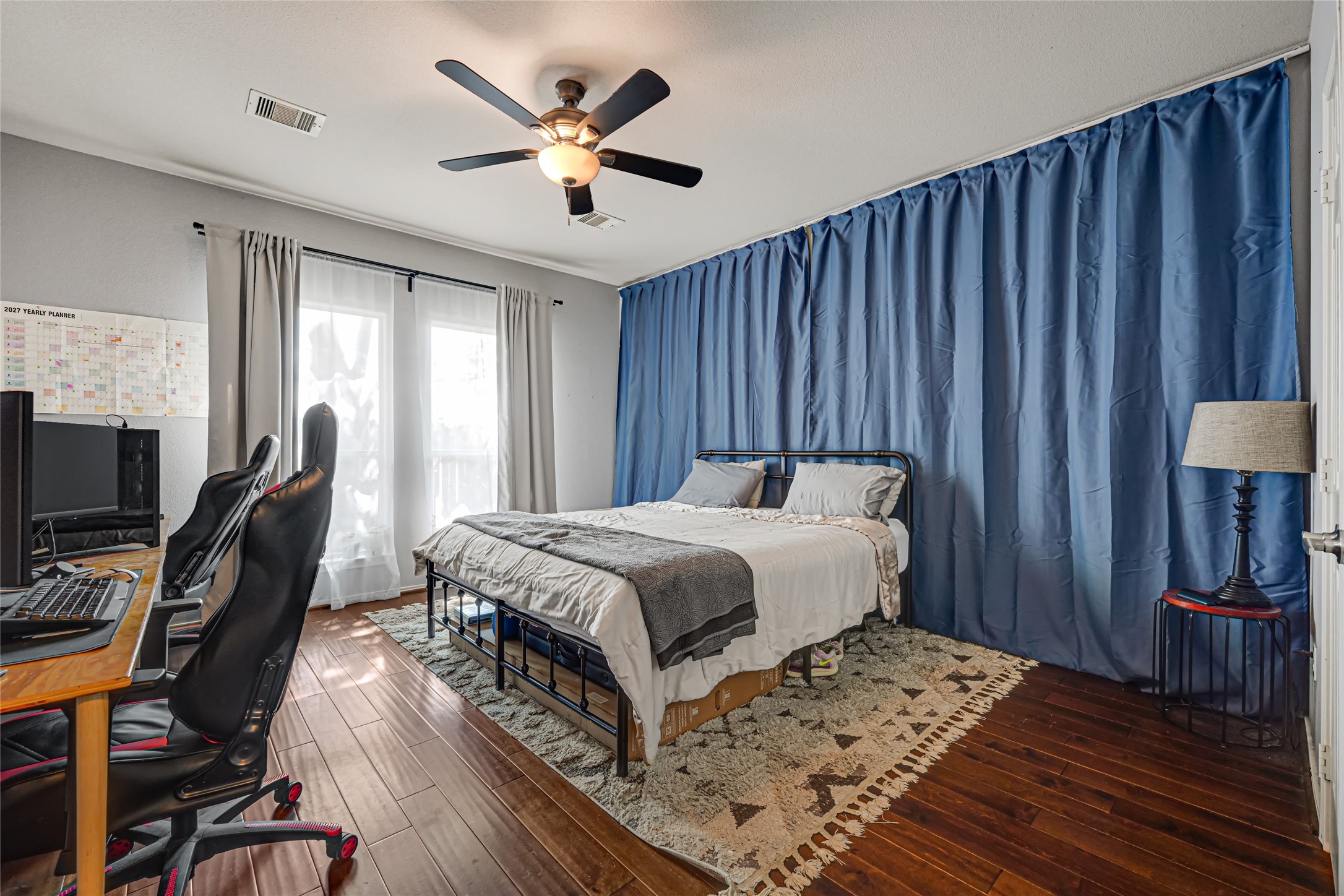 Spacious bedroom with rich hardwood floors, featuring a cozy bed, stylish blue curtains, and a practical workspace area. Spacious bedroom with rich hardwood floors, featuring a cozy bed, stylish blue curtains, and a practical workspace area.