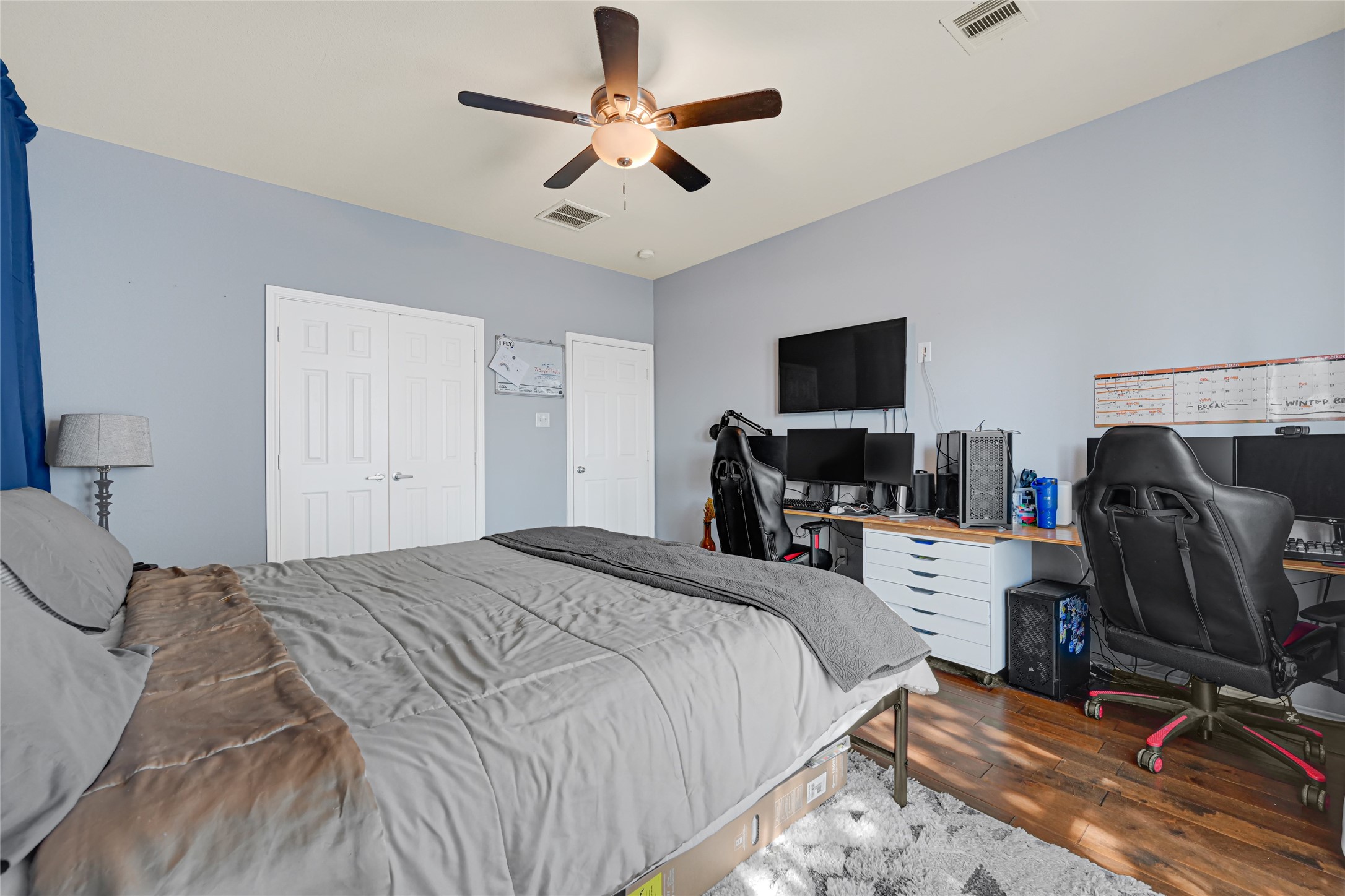 Spacious bedroom featuring a comfortable bed, dual workspace with mounted monitors, and ample storage in a serene, light-filled setting.