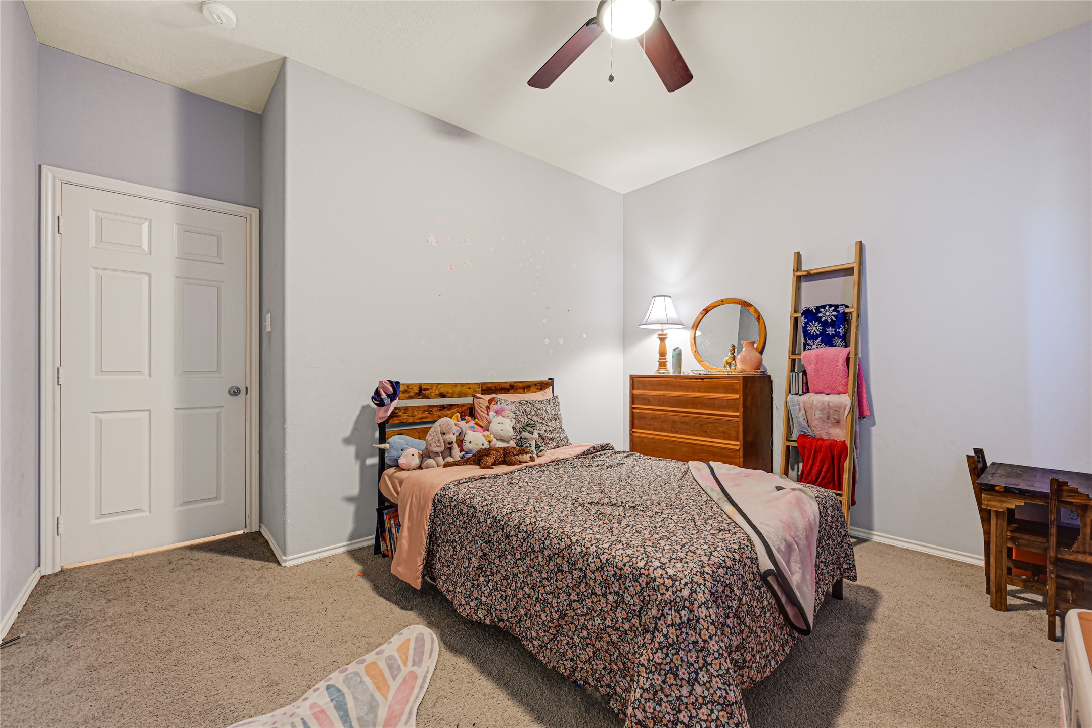 Cozy bedroom featuring soft carpeting, a charming floral bedspread, and a ceiling fan for comfort. Natural light highlights the wooden dresser and decorative ladder, creating a warm, inviting atmosphere.