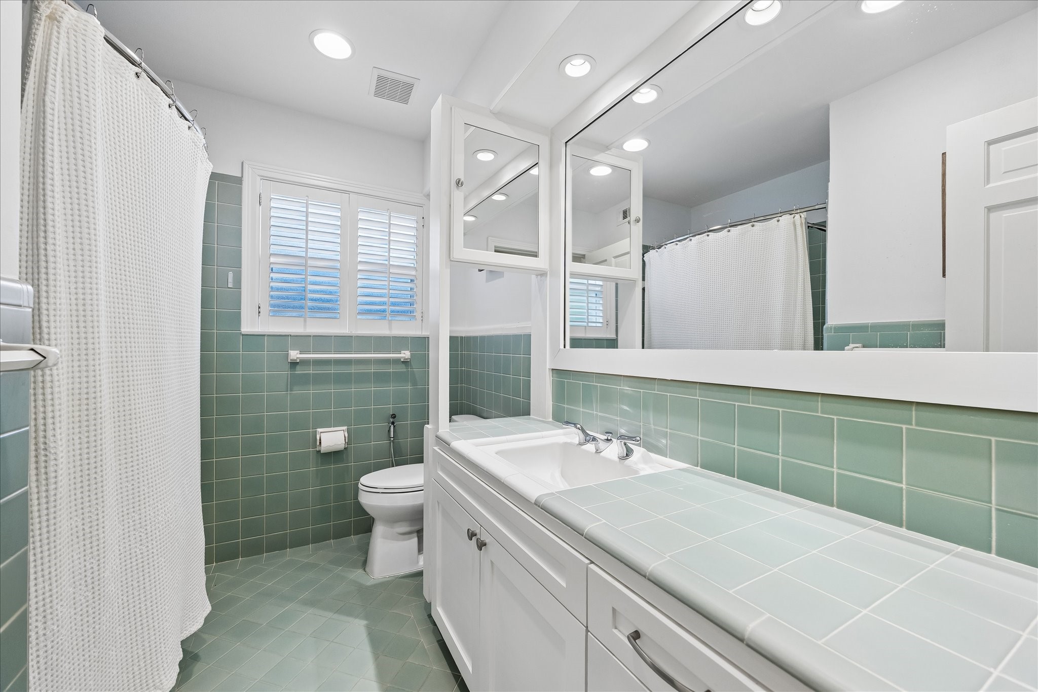 Secondary bath features classic mint-green tile flooring, walls & countertop, white cabinetry w/storage, oversized vanity mirror, recessed lighting & tub/shower combination.
