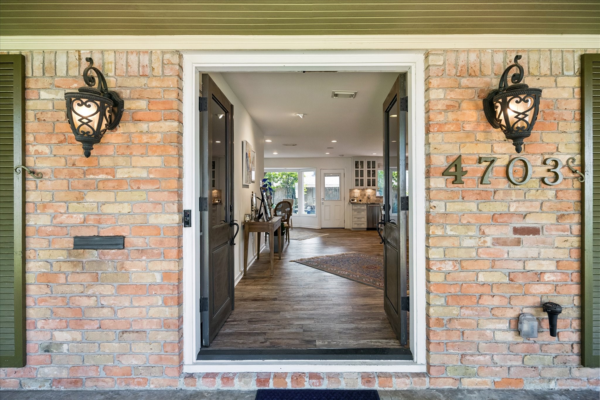 Welcoming double-door entry framed by brick accents & decorative lanterns opens to an inviting interior w/wood flooring & views into the living spaces beyond.