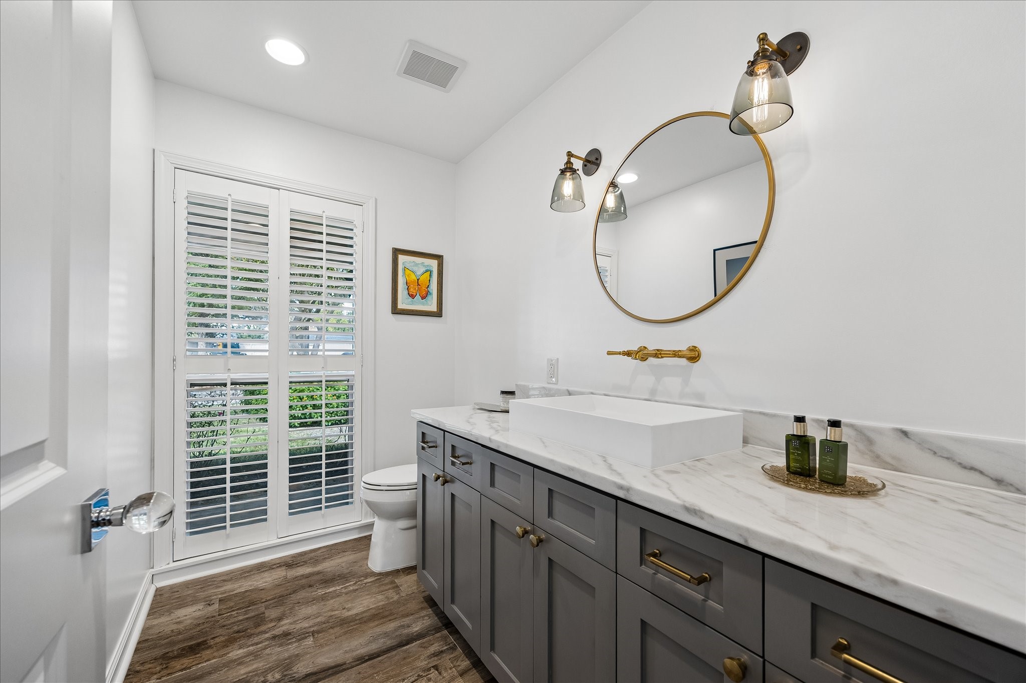 Beautiful half bath features a sleek rectangular vessel sink, stone countertop, gray cabinetry w/brass hardware & a stylish round mirror. Decorative wall sconces, wall-mounted faucet, wood flooring & large window w/plantation shutters complete the space.