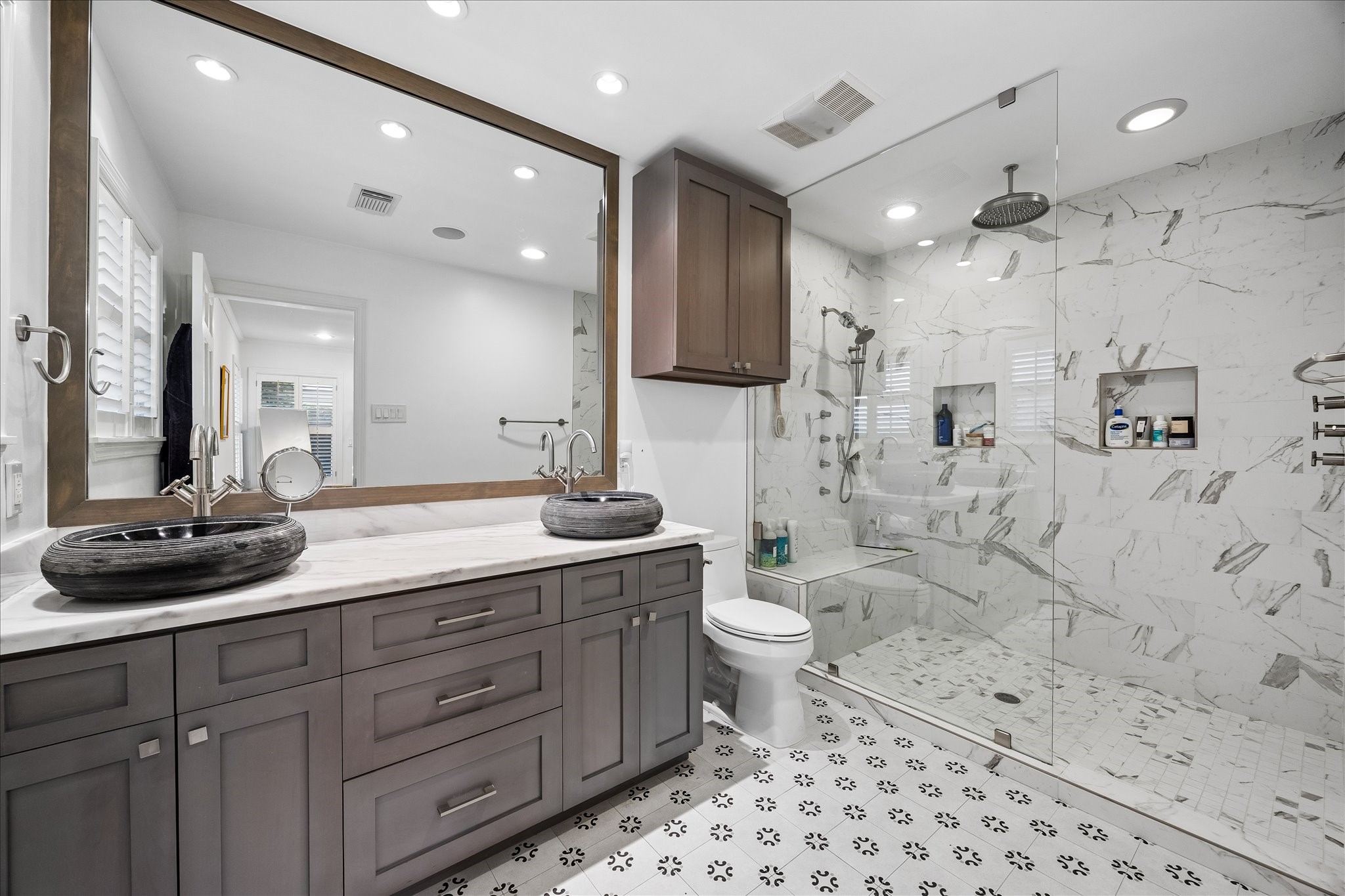 Beautifully updated primary bath features dual vessel sinks, stone countertop, custom cabinetry, oversized mirror & decorative tile flooring. Expansive glass-enclosed shower offers gorgeous tile, rainfall showerhead, handheld shower, built-in bench & recessed niches.