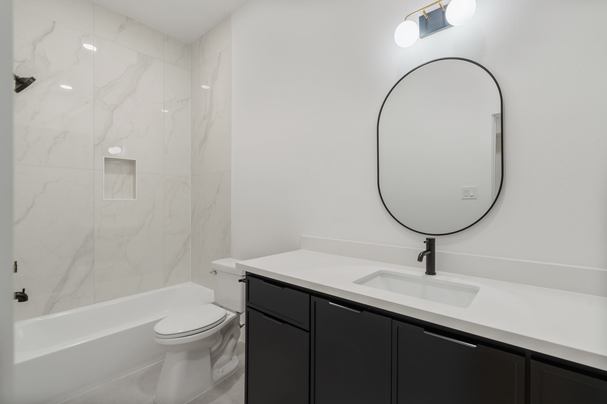 Elegant bathroom featuring a sleek black vanity, modern fixtures, and a stylish oval mirror, complemented by a pristine white tub and marble-patterned tiles for a contemporary feel.