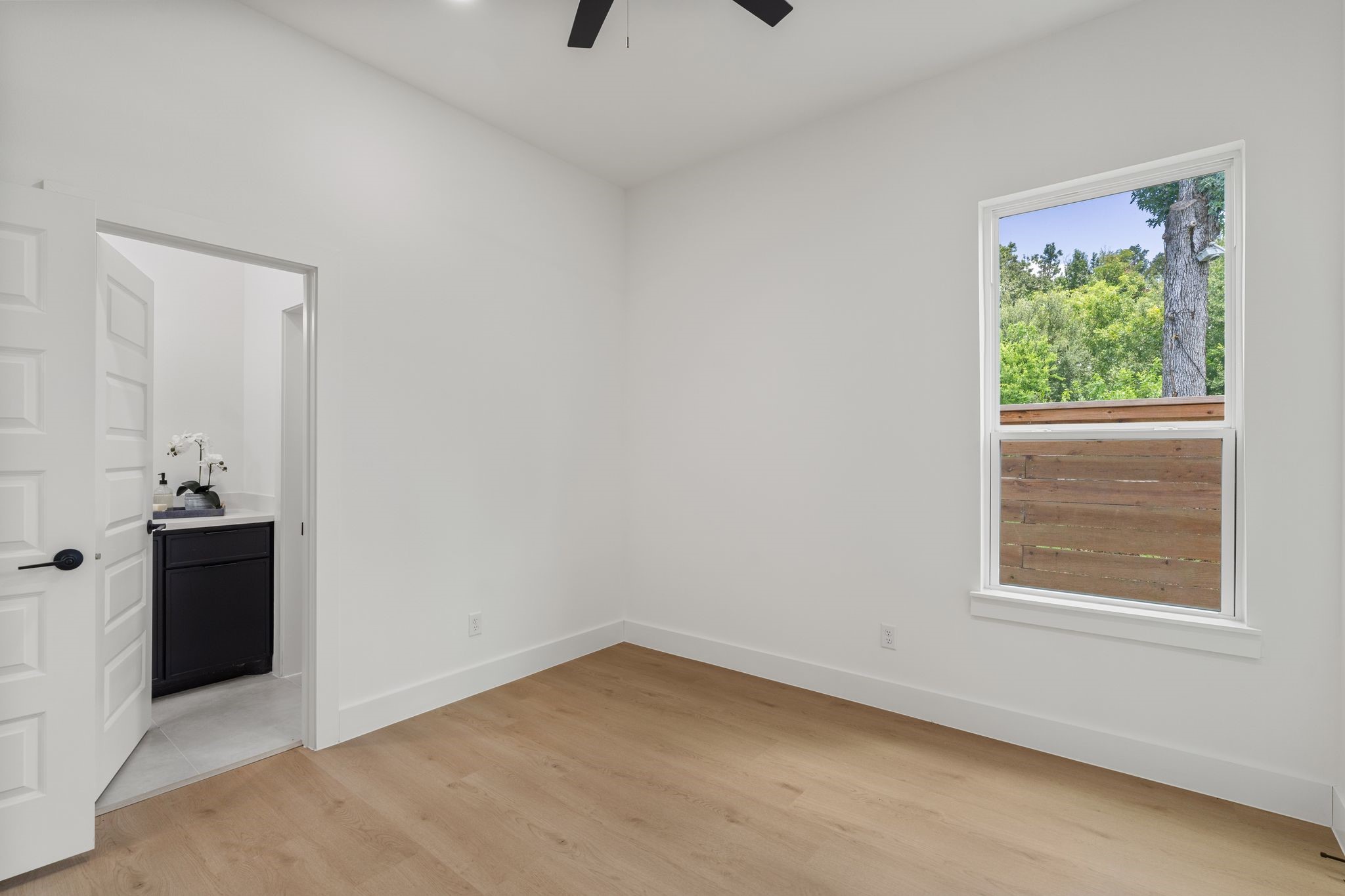 Bright, airy room with sleek hardwood floors and amodern ceiling fan, featuring a window with lush green views and an adjacent bathroom with contemporary fixtures.