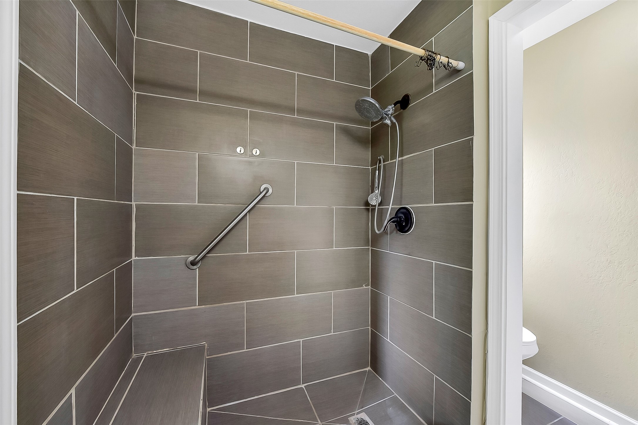 The master bathroom features a modern design with a sleek vanity topped with a solid-surface countertop, framed mirror, and updated lighting. The spacious walk-in shower is accented with oversized tile, a built-in bench, a grab bar, and dual showerheads, including a rain-style fixture.