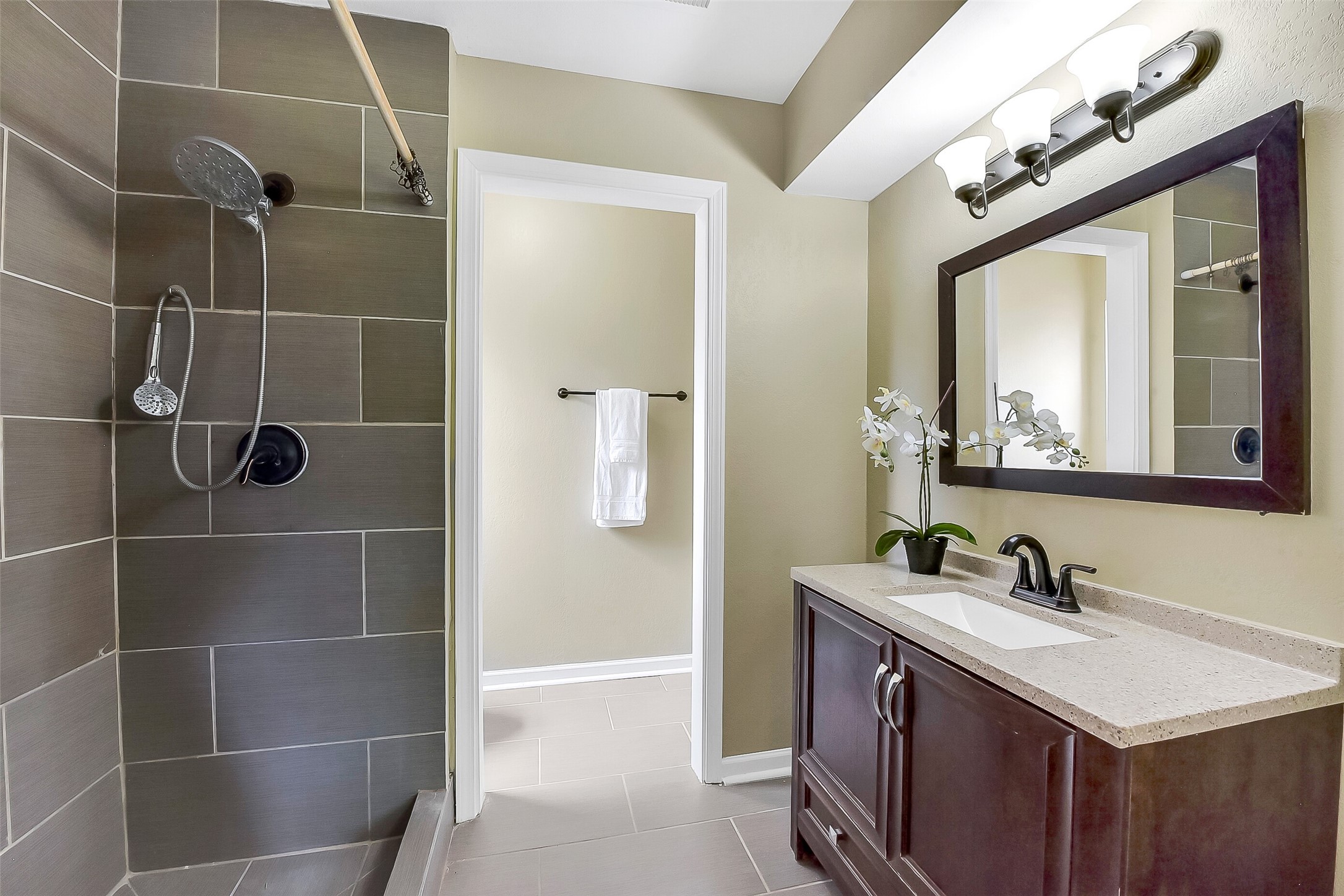 The master bathroom features a modern design with a sleek vanity topped with a solid-surface countertop, framed mirror, and updated lighting. The spacious walk-in shower is accented with oversized tile, a built-in bench, a grab bar, and dual showerheads, including a rain-style fixture.