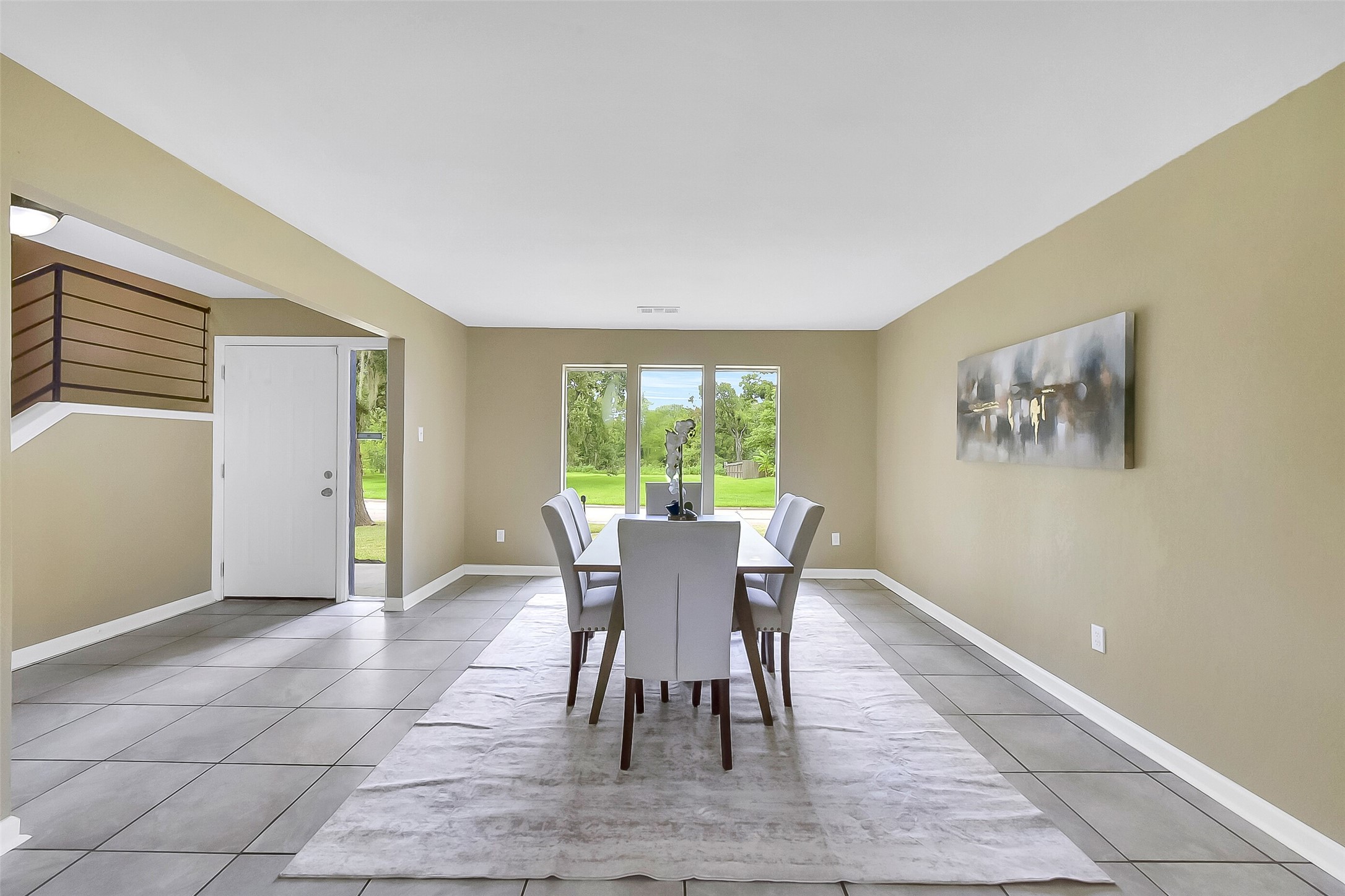 Formal dining room with neutral paint, large-format tile floors, and abundant natural light from a trio of wide picture windows overlooking the front yard.