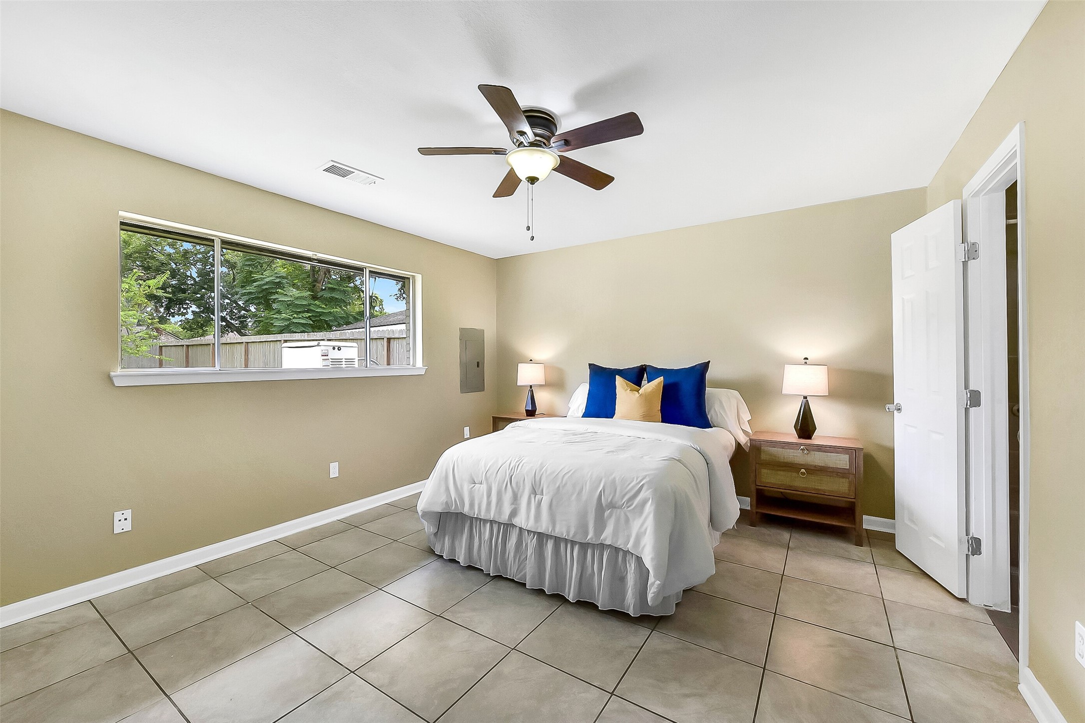 This first-floor primary suite offers a peaceful retreat with neutral tones, tile flooring, a ceiling fan, and a large window that fills the room with natural light. An en-suite bath provides added privacy and convenience.
