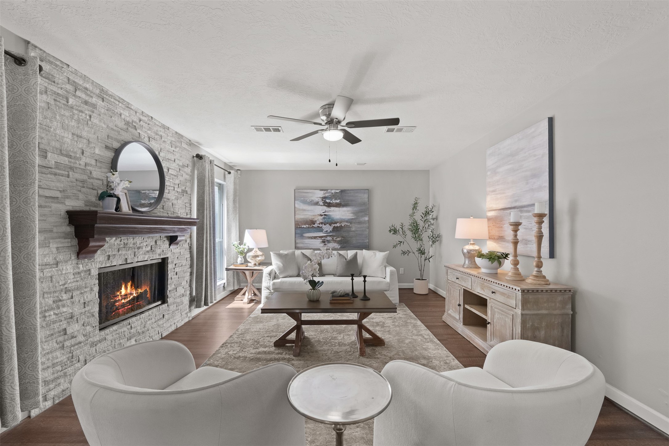 A floor-to-ceiling stone stack fireplace anchors this inviting living space, accented by warm wood flooring and a classic ceiling fan. Soaring natural light streams in from surrounding windows, highlighting an open layout ideal for relaxed hosting.
