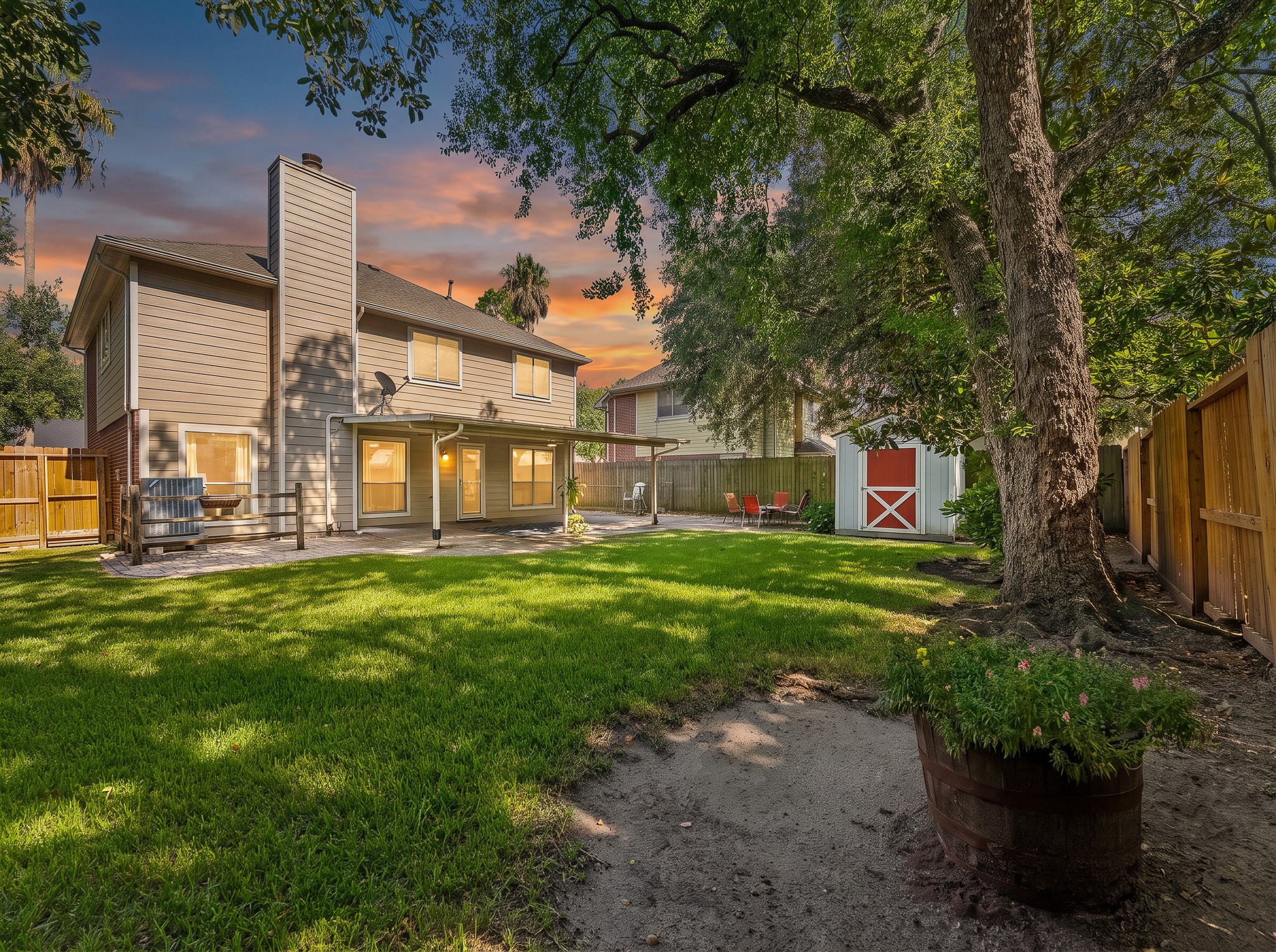 Shaded by magnificent trees, this expansive grassy backyard features a covered patio, paved seating area, full privacy fencing, and a distinctive red storage shed.