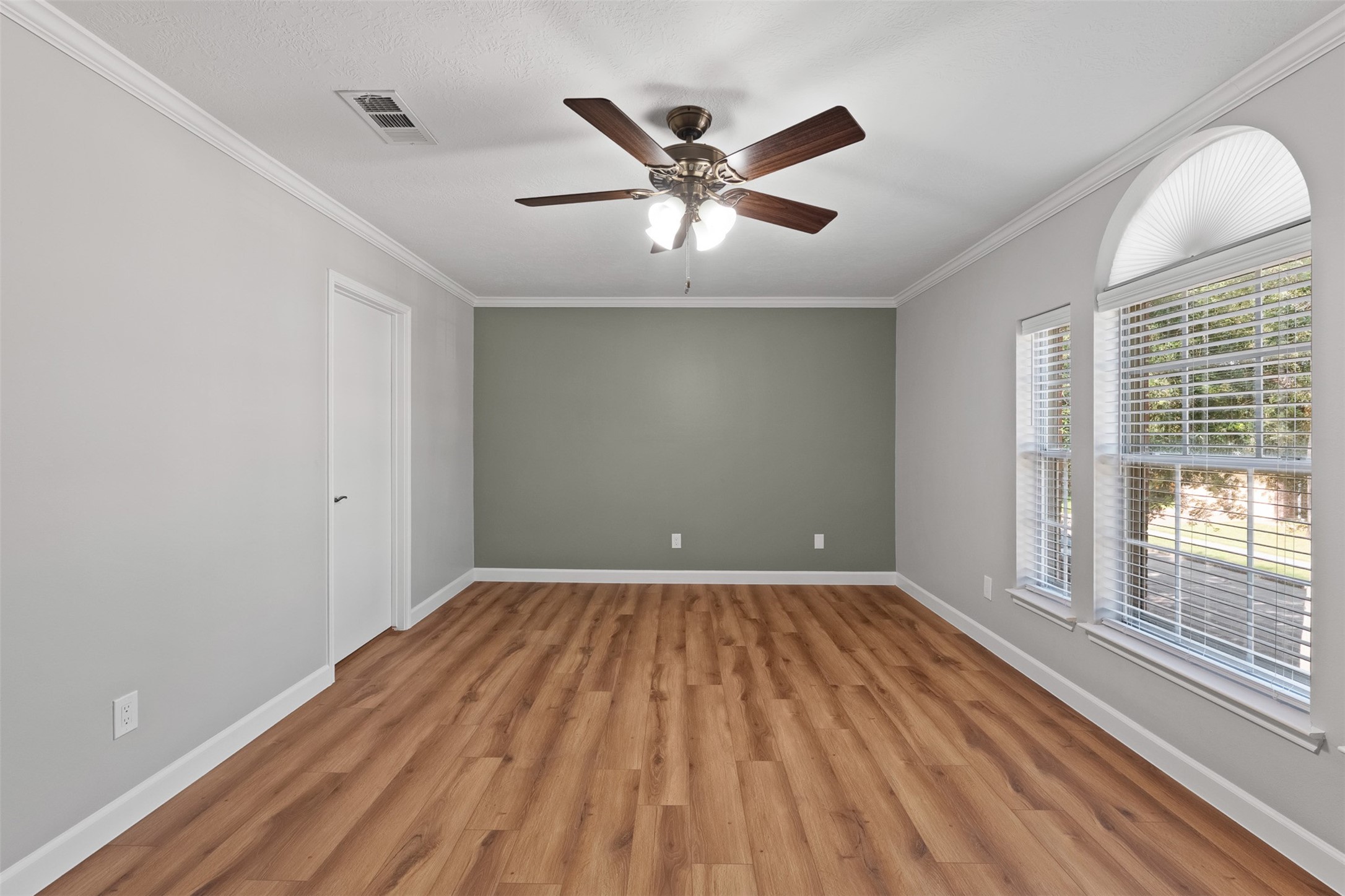A sage green accent wall complements the warm wood-look flooring and crisp crown molding in this secondary bedroom. Wide windows topped with a decorative arch bring in abundant light, while a dark wood ceiling fan adds style and breeze.