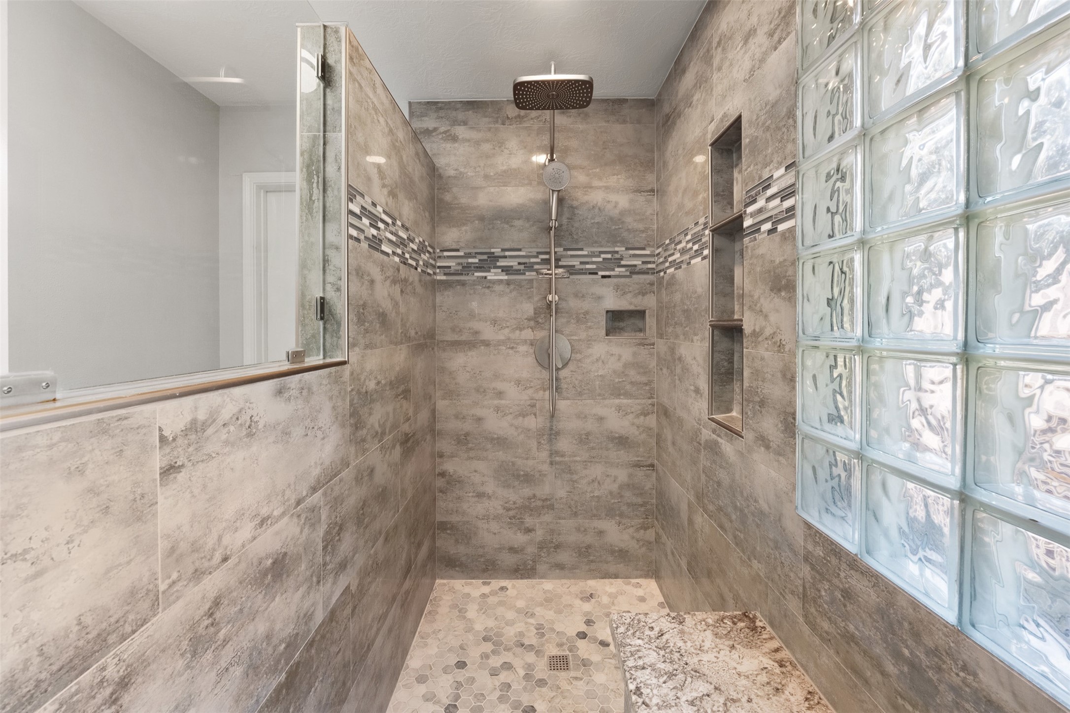 Lined with large-format grey tile, this renovated shower features a rain shower head, glass block window, built-in bench, and hexagon tile floor with a mosaic accent band.