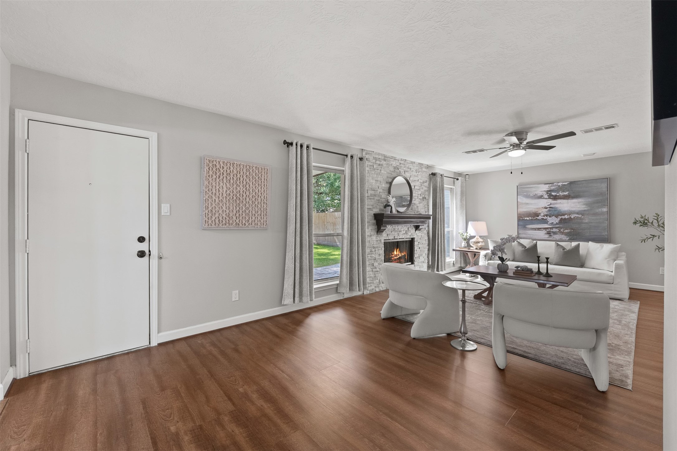 Rich wood-look flooring leads directly into a light-filled living area centered around a floor-to-ceiling stacked stone fireplace. Open-concept sightlines extend toward the backyard, complemented by neutral walls, modern ceiling fan fixtures, and direct access through the entry door.