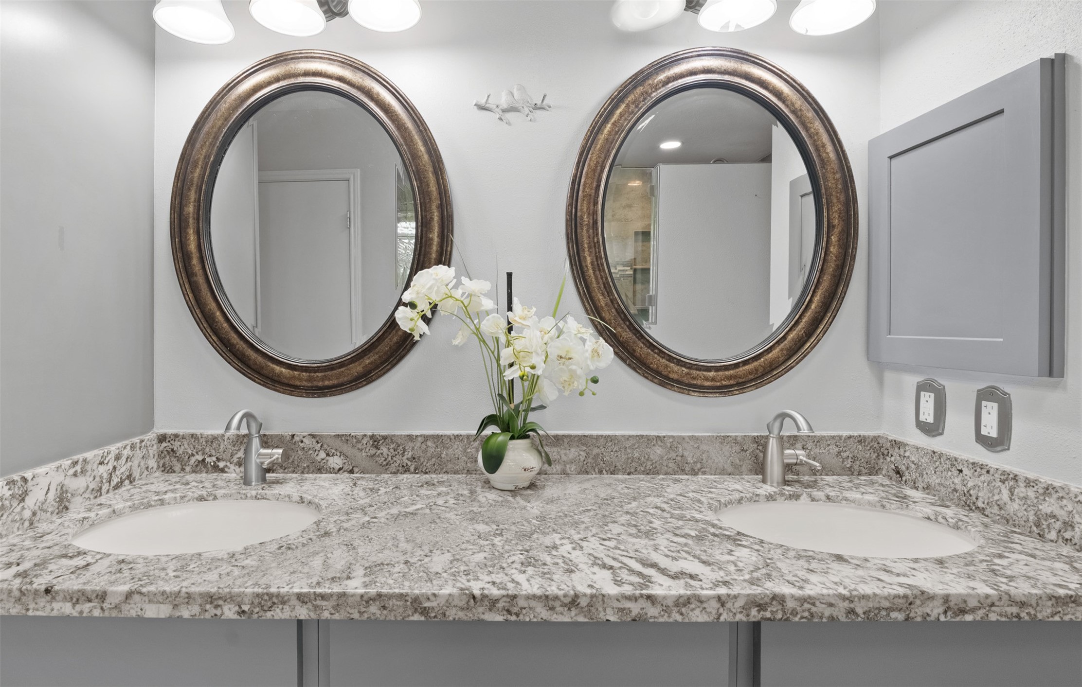 Renovated vanity with granite countertops features a dual sink layout with brushed nickel faucets and oval mirrors. Custom grey wall-mounted storage and updated vanity lighting complete this spa-inspired space.