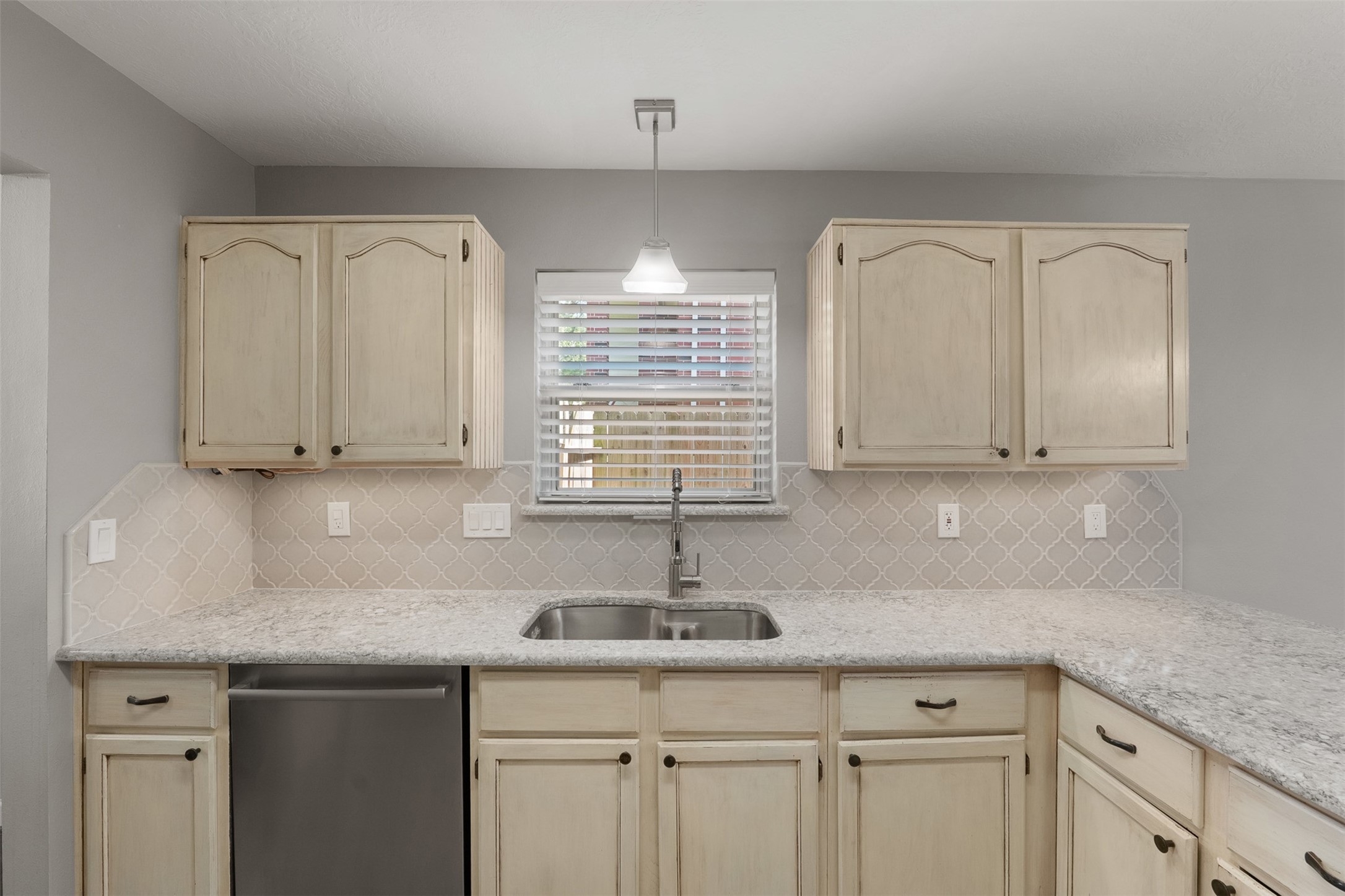 Quartz countertops, a intricate designer backsplash, and a spacious dual sink anchored under a window refine this culinary space. Off-white cabinetry offers ample storage, complimented by sleek stainless steel appliances and a modern pendant light.